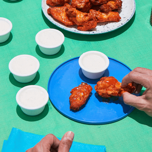 Person holding a piece of fried chicken with hot sauce on a blue plate, surrounded by small cups of ranch dressing and more fried chicken on a white plate, on a green table.