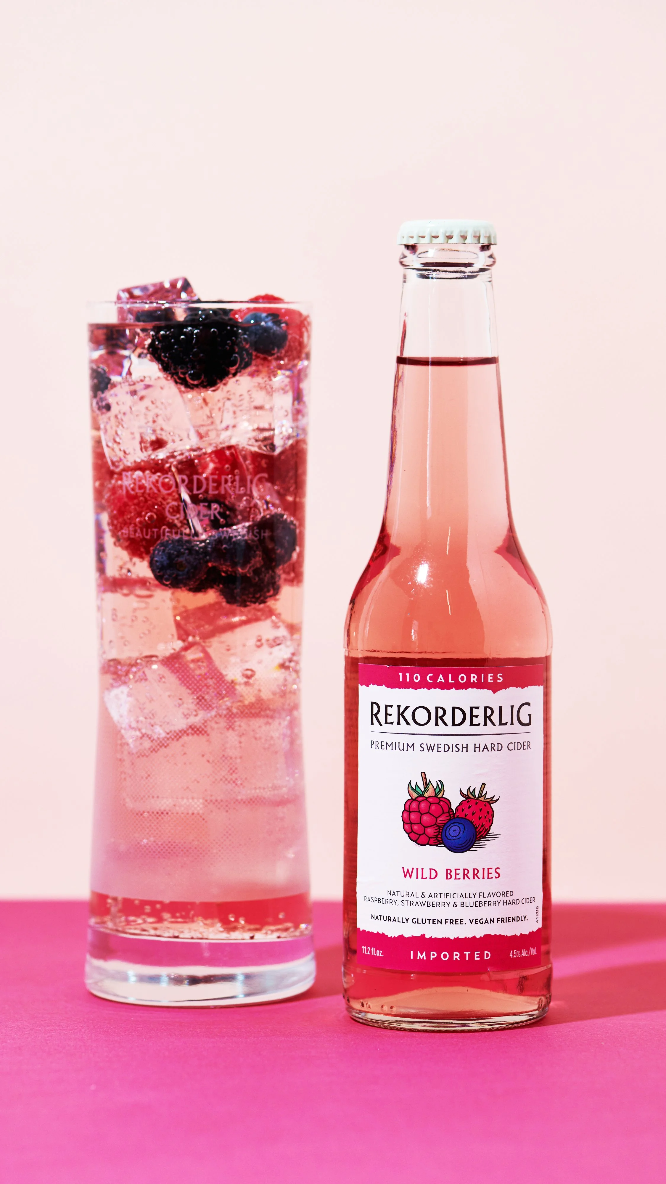 A glass of pink sparkling beverage with blackberries and ice next to a pink bottle of Rekorderlig Wild Berries cider.