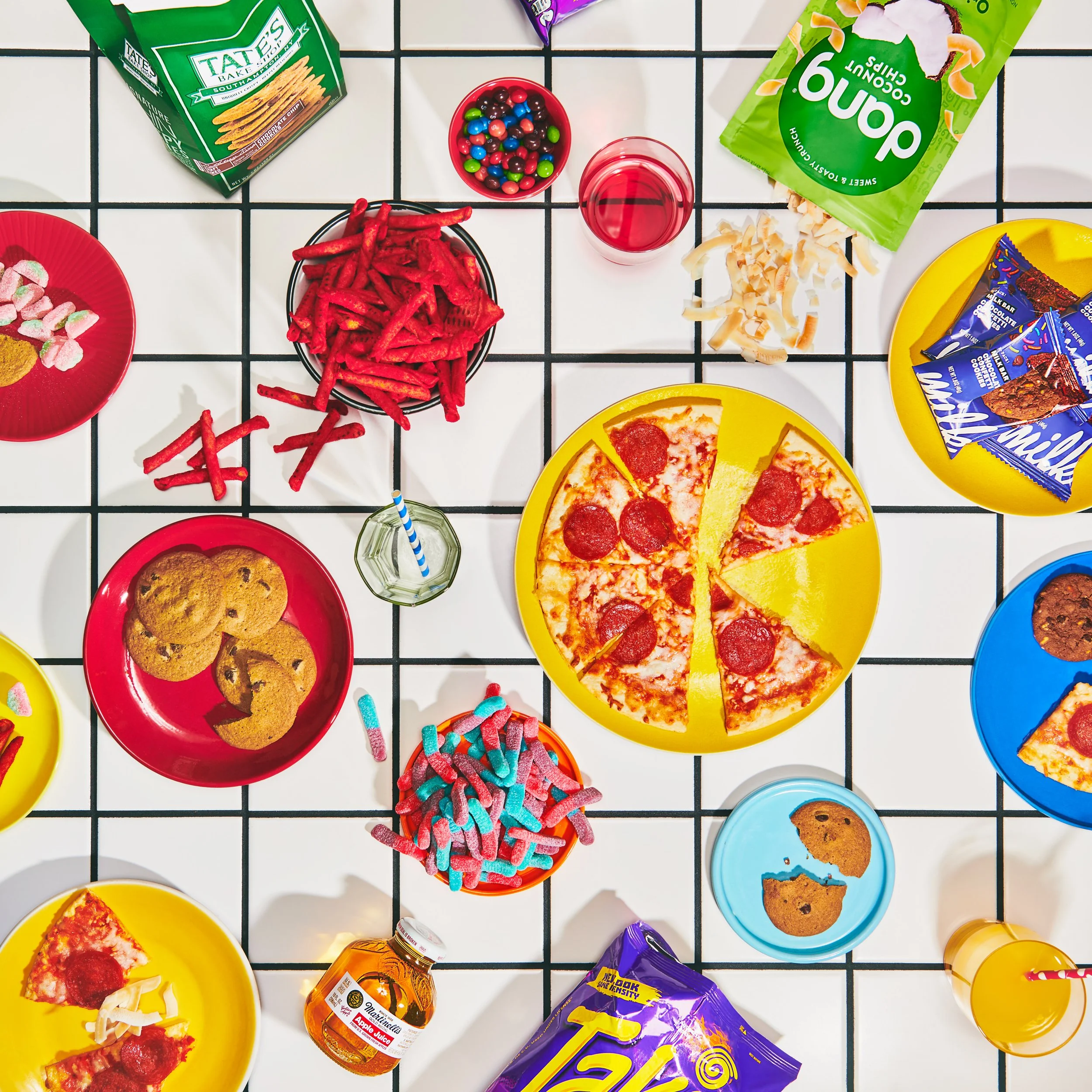 A table set for a snack with various food items including a pepperoni pizza, cookies, gummy worms, cotton candy, chips, candy bars, and drinks, on a white tiled surface.