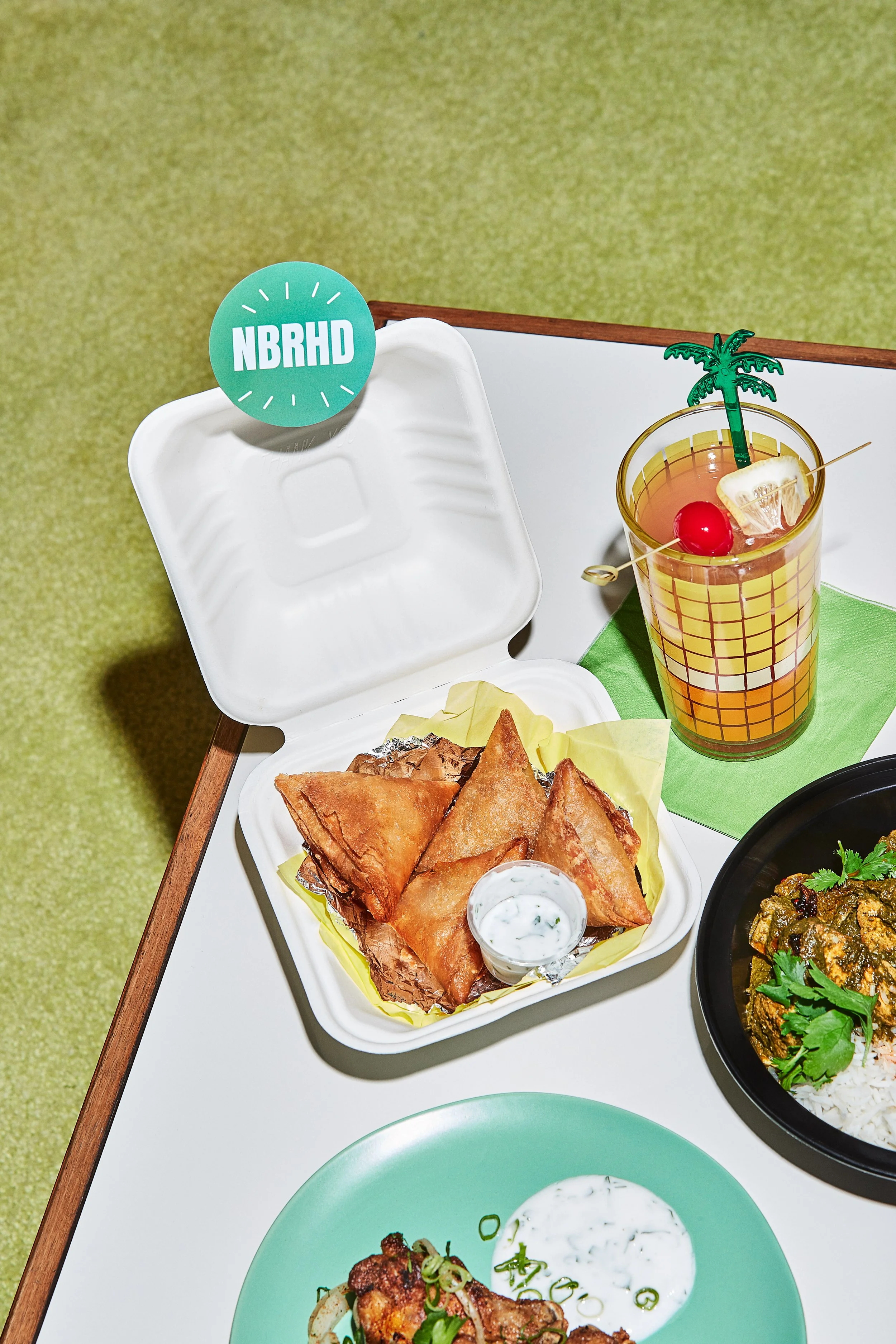 A takeout container with samosas, a small cup of yogurt sauce, a colorful drink with a cherry and lemon slice, and a plate of curry with rice on a table.
