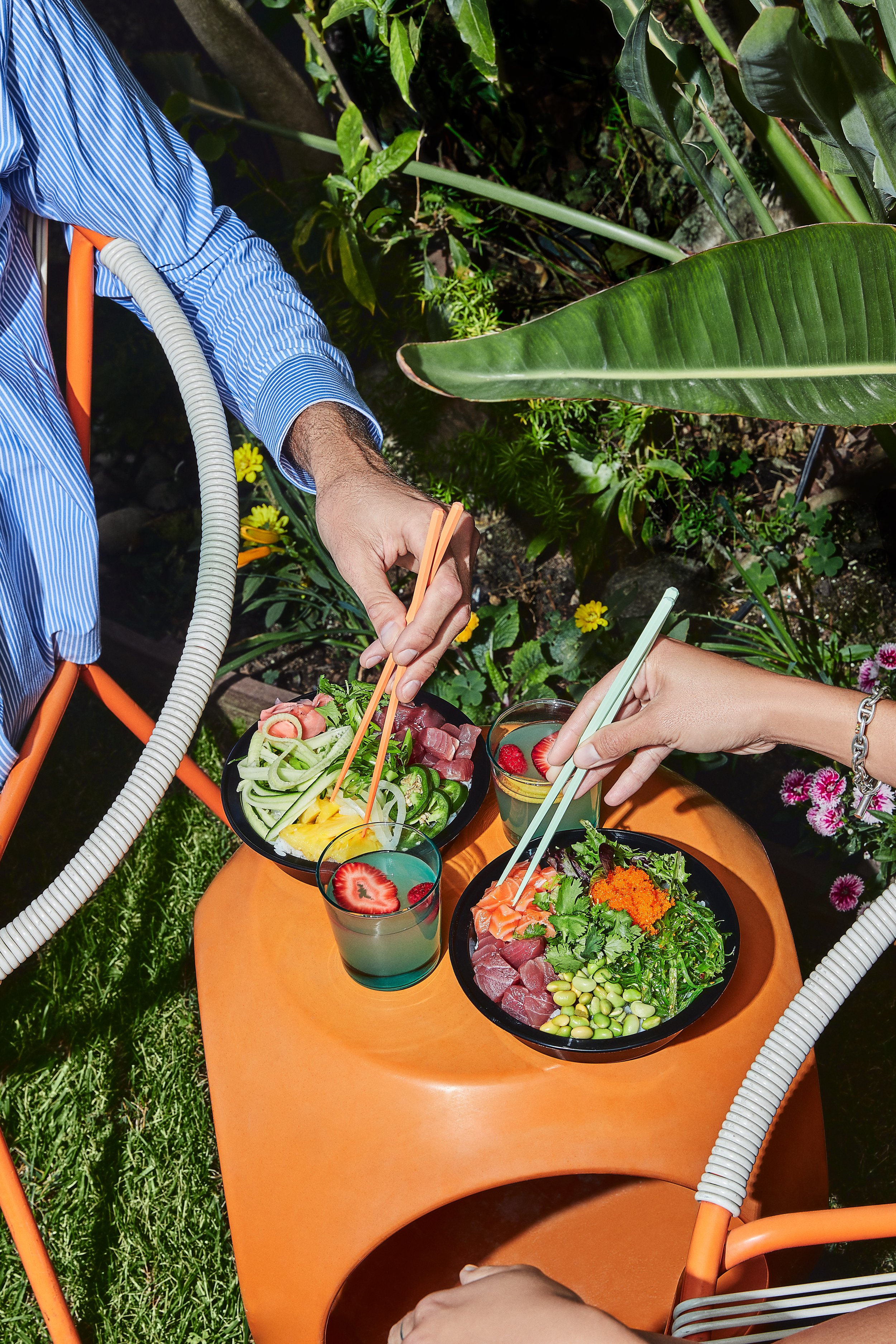 Two people are having a meal outdoors at a small orange table, with bowls of sushi and drinks. One person is picking up food with chopsticks, and the other has a glass drink with fruit slices.