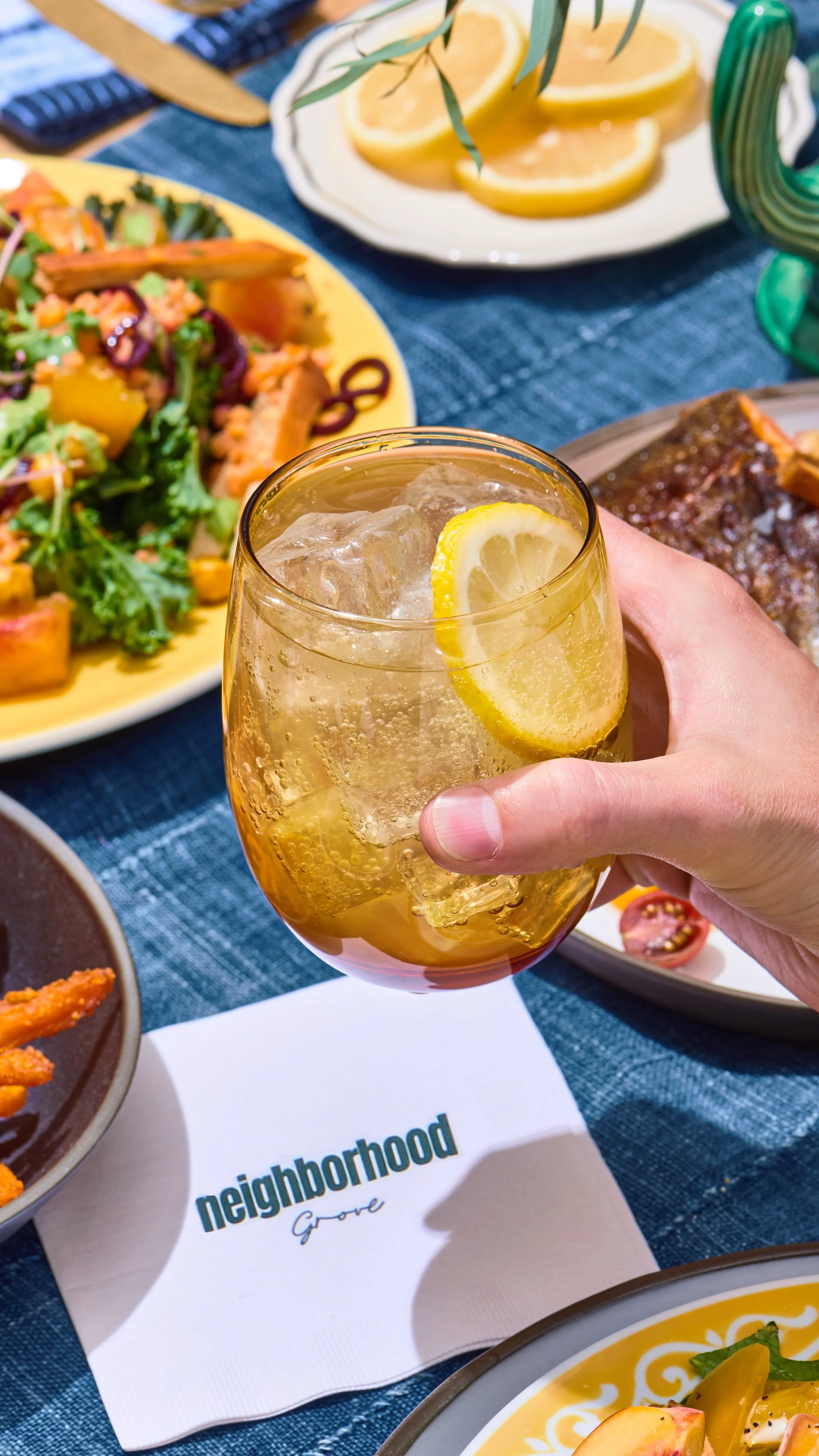 A glass of iced lemon soda held by a person at a restaurant table with various dishes, including a salad, lemon slices, a glazed meat dish, and fried food, on a blue tablecloth.