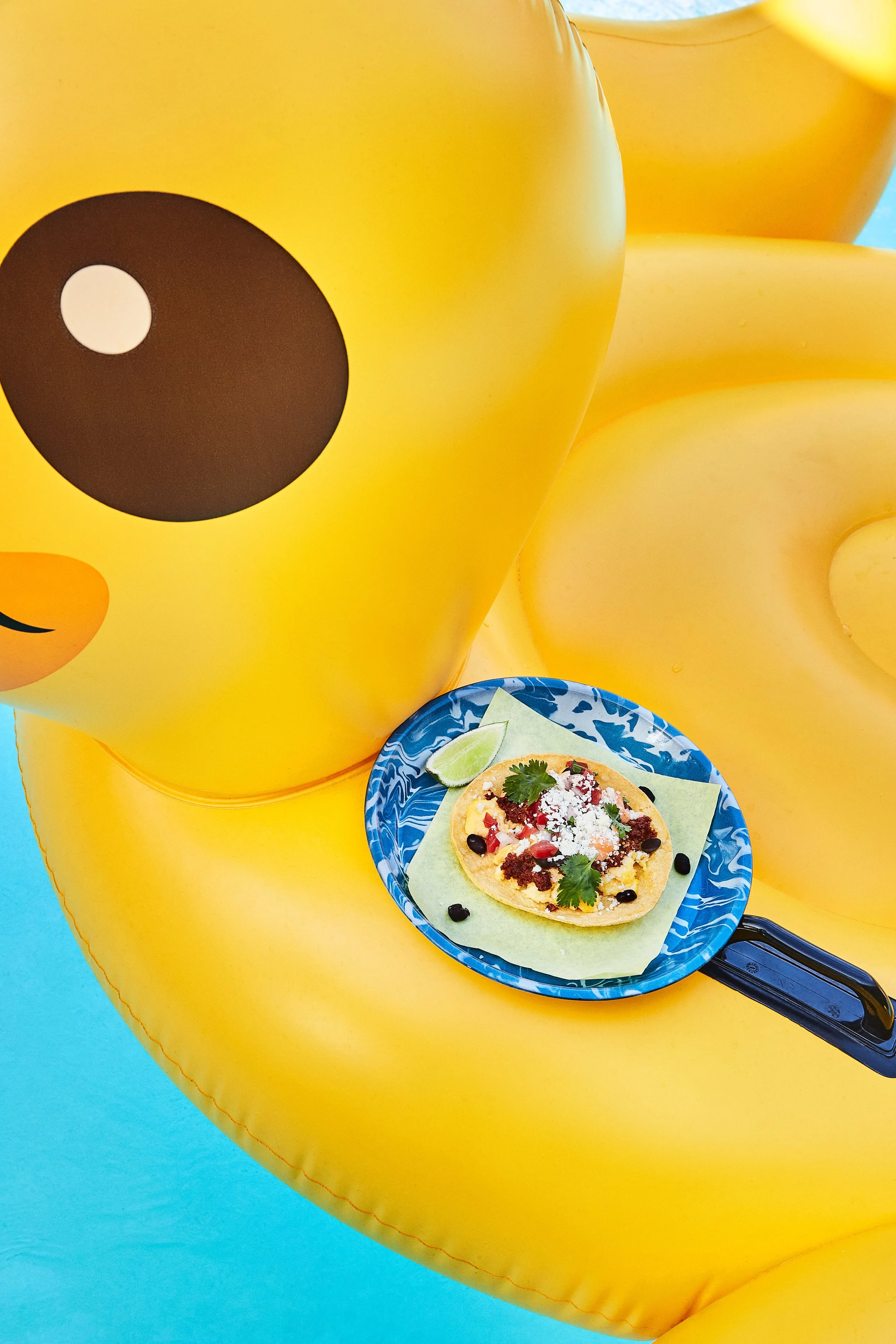A close-up image of a large yellow inflatable rubber duck with a black eye, holding a blue plate with a taco, lime wedge, and garnishes, against a yellow and blue background. food and drink shoot, editorial, commercial 