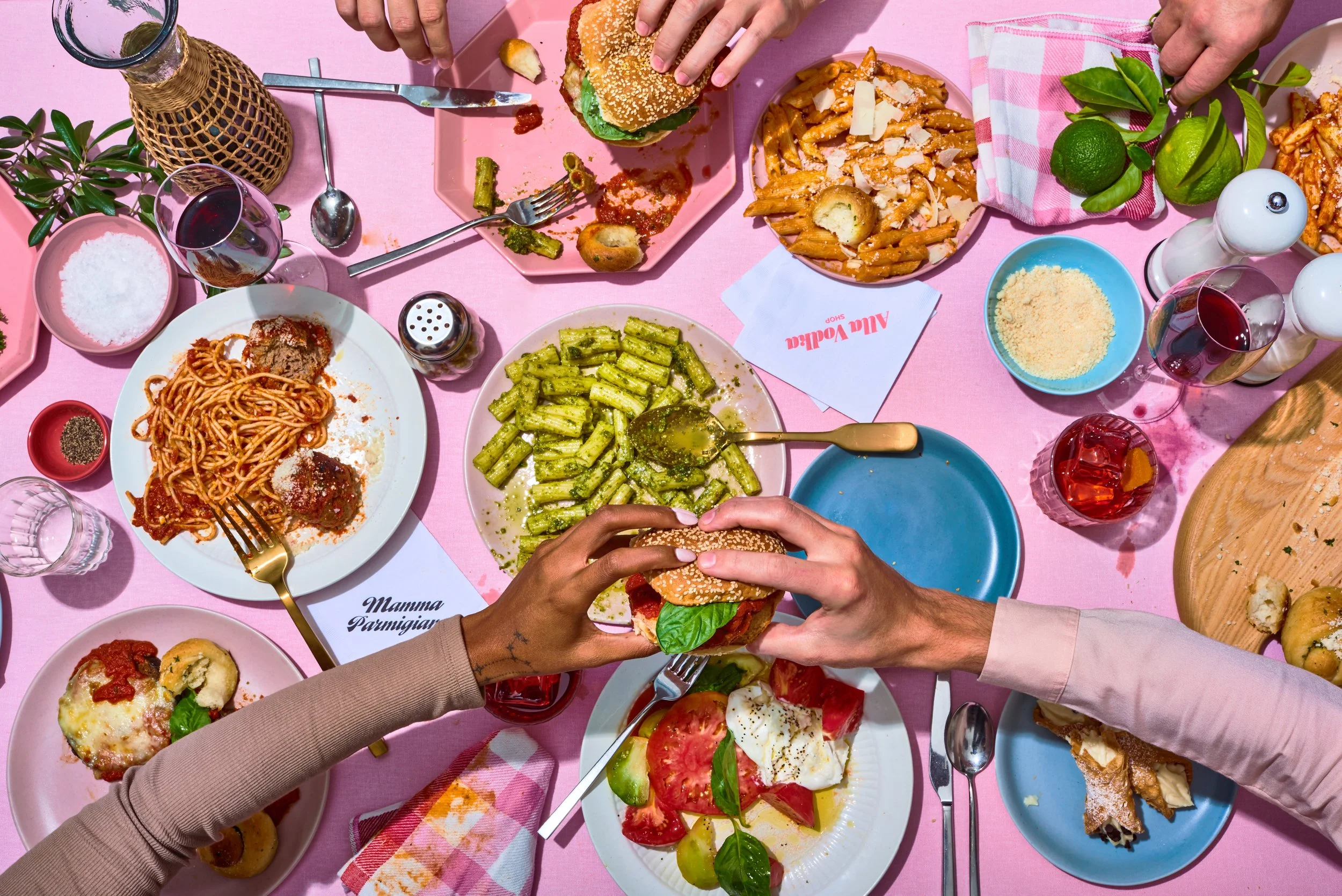 A pink table filled with various Italian dishes, drinks, and condiments, with two people holding a burger together.