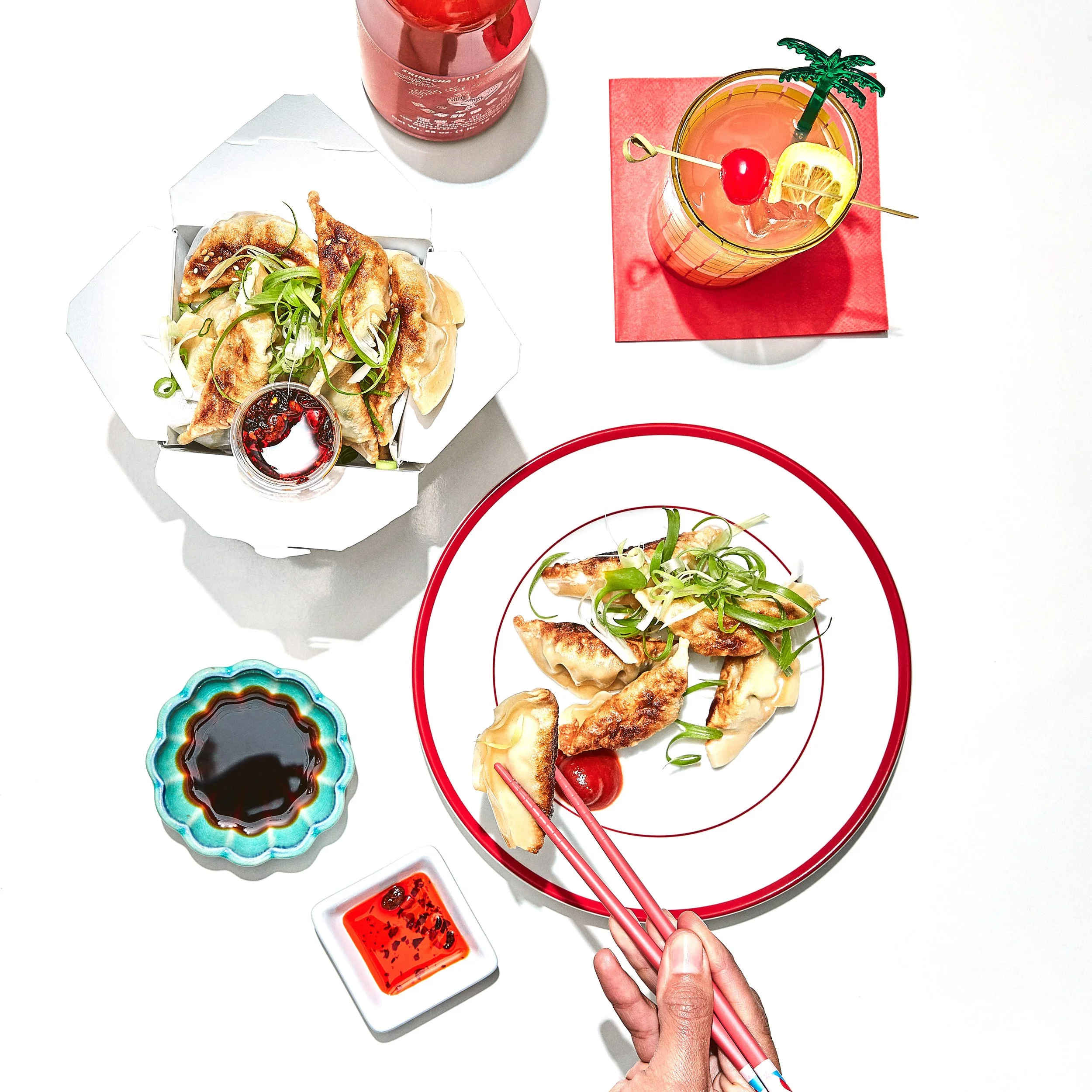 Top-down view of a table with Asian cuisine, including a plate of fried dumplings with green onions, a bamboo steamer basket of dumplings, a small bowl of soy sauce, a glass of pink cocktail with a cherry and lemon garnish, and a can of soda.