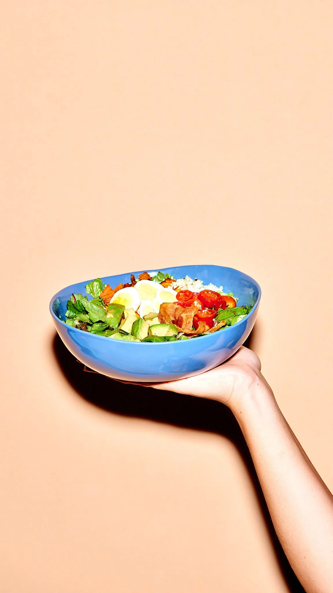 A hand holding a blue bowl filled with salad including lettuce, cherry tomatoes, boiled eggs, cucumber, and bacon against a beige background.