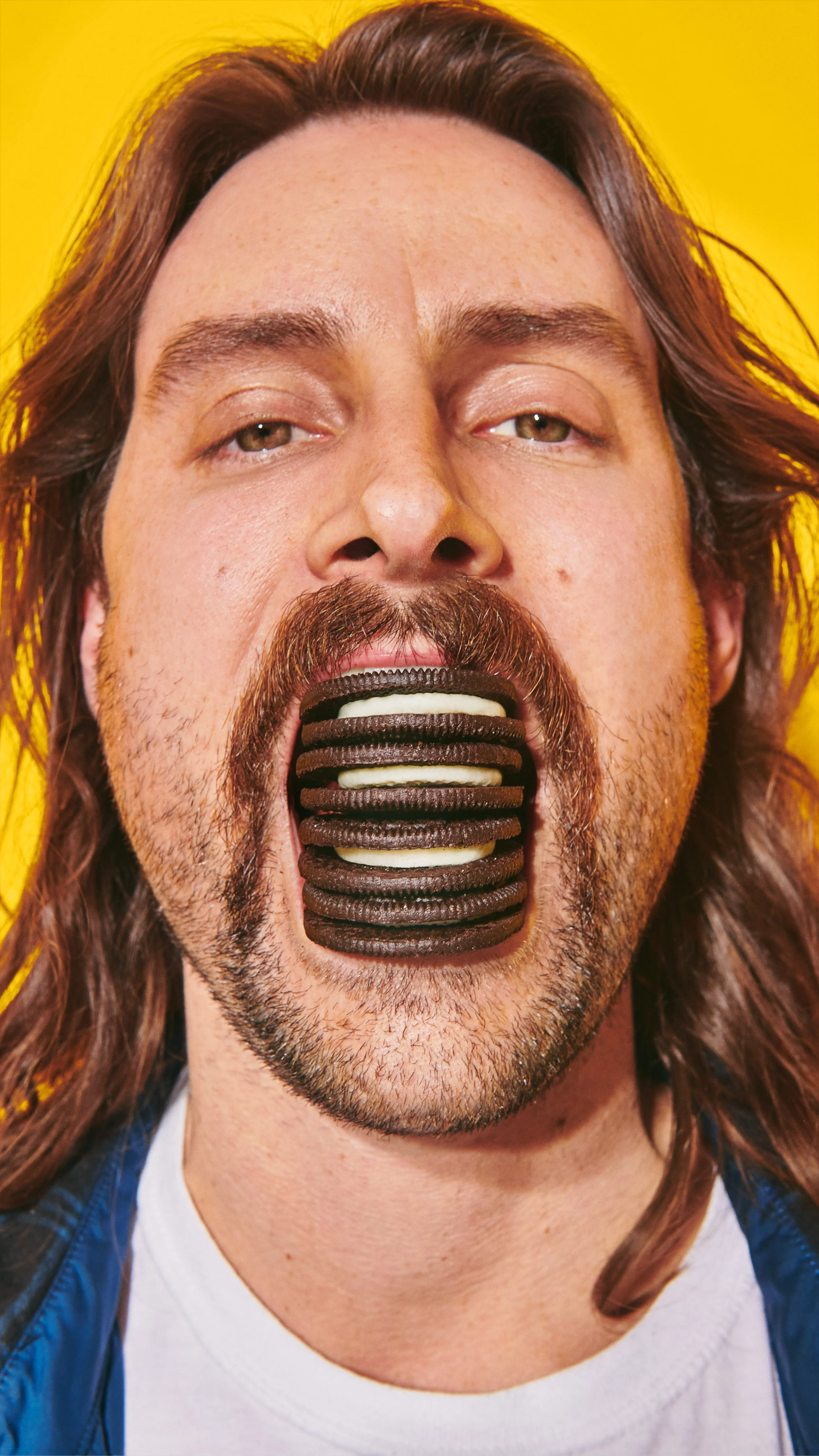 A man with long hair and a beard has his mouth open, holding layered Oreos in his mouth, against a yellow background.