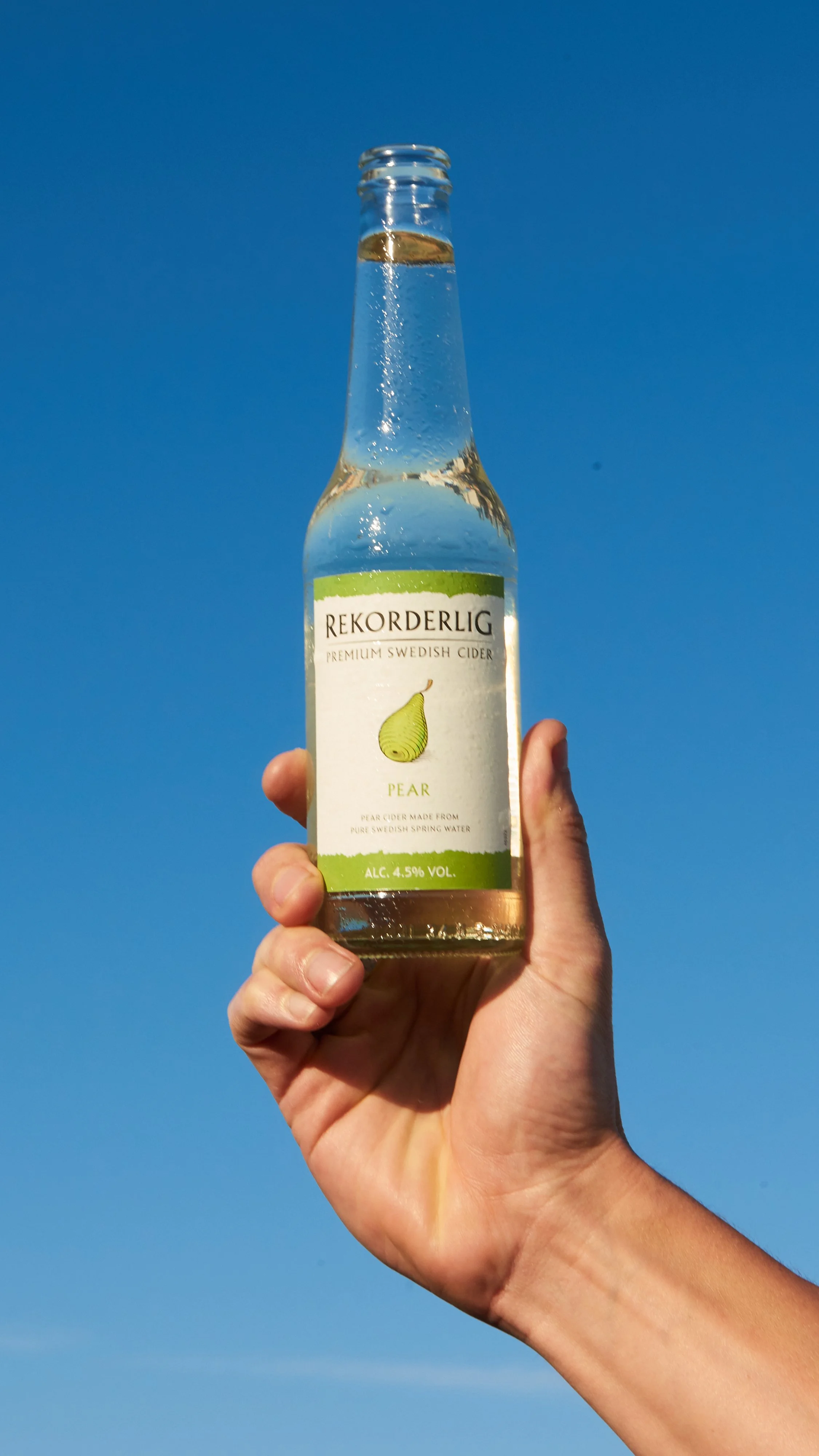 A hand holding a bottle of Rekorderlig pear Swedish cider against a clear blue sky.