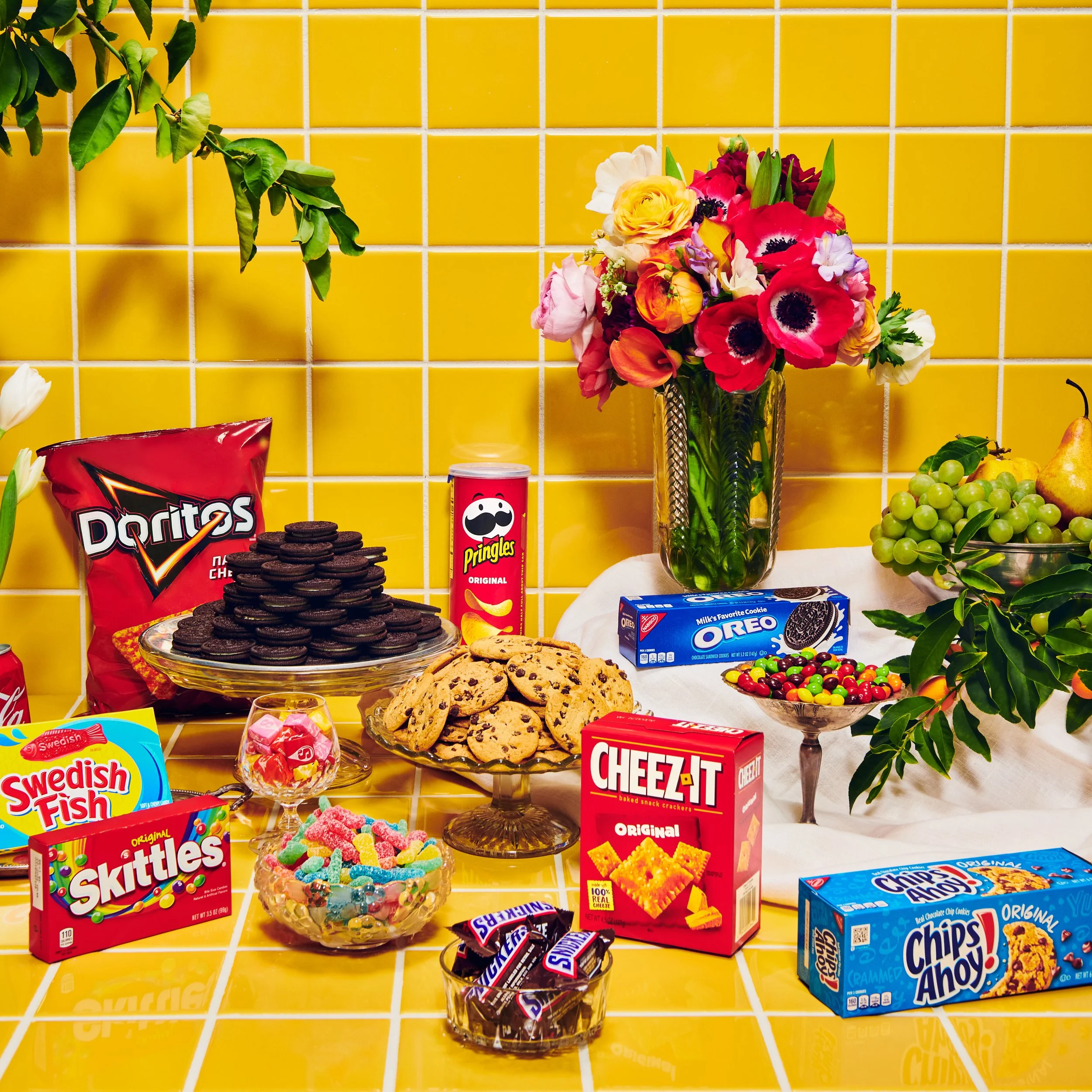 Assorted snacks and candies on a yellow tiled table, with a bouquet of multicolored flowers in a glass vase against a yellow tiled wall.