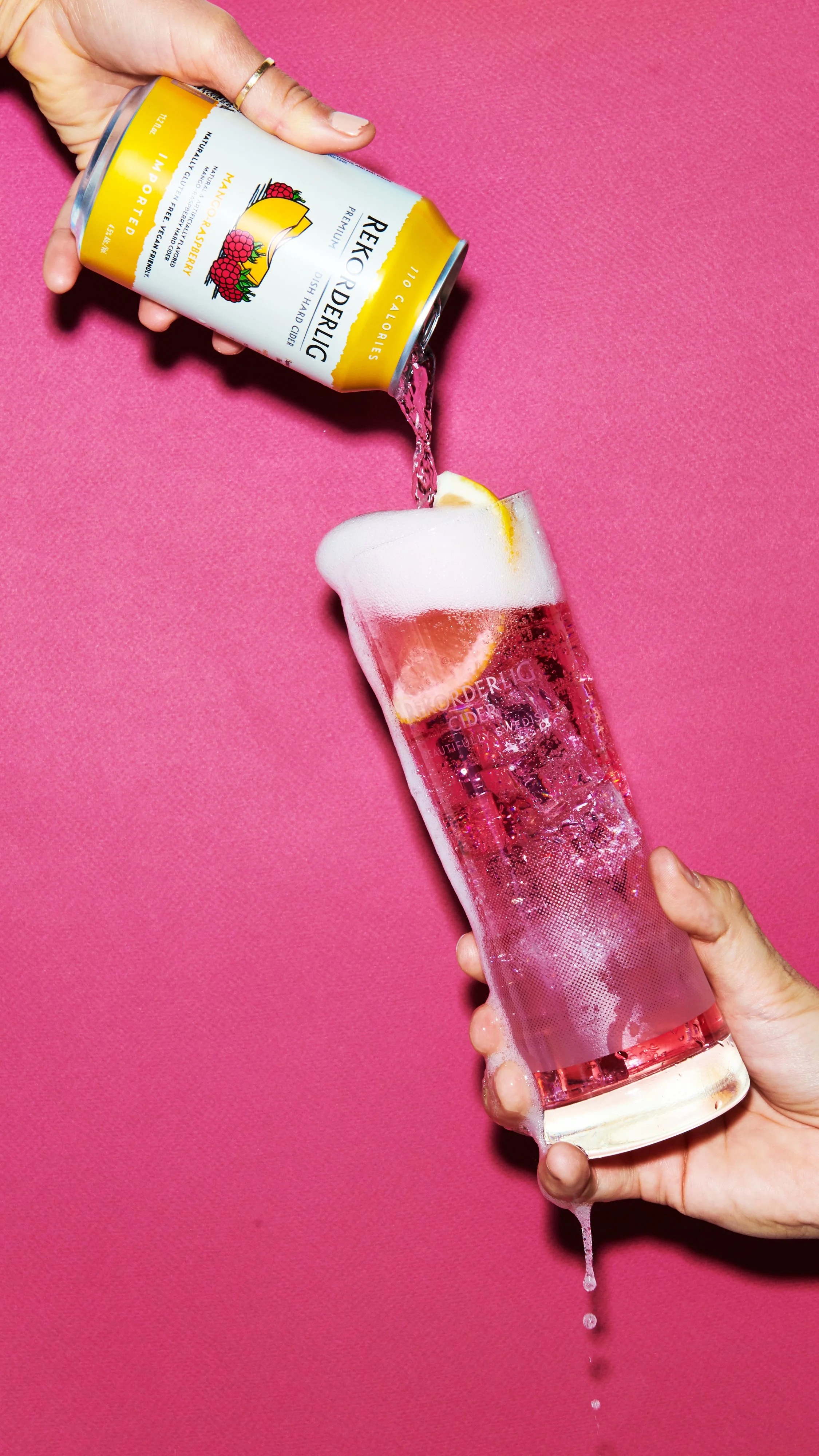 A person pouring pink sparkling cider into a glass with lemon slices against a pink background.