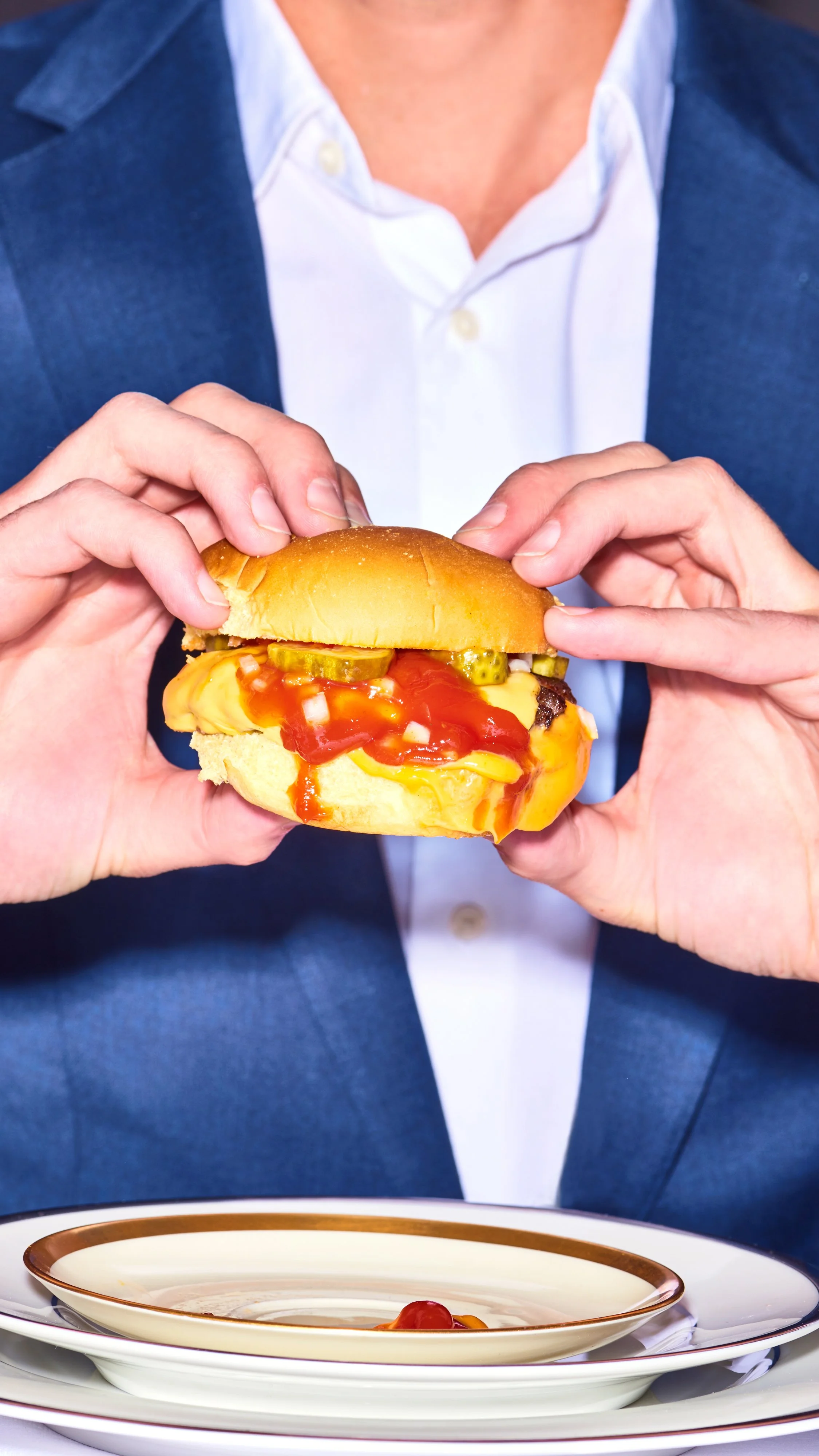 Person in a blue blazer holding a cheeseburger with ketchup and pickles, standing over a stack of dishes.