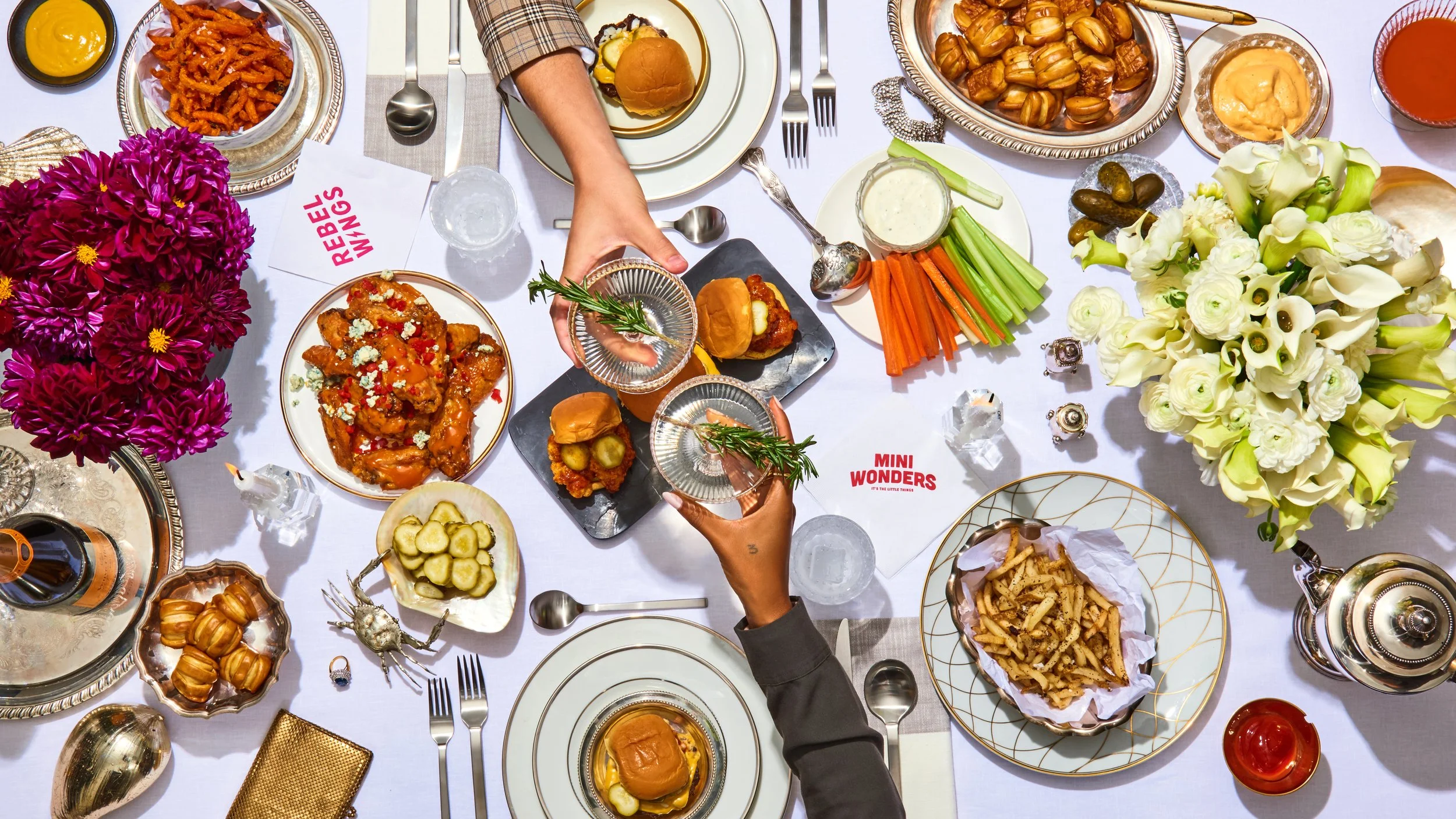 A colorful table setting with various foods, drinks, and flowers, including fried chicken, pasta, vegetables, and desserts, with two people clinking glasses.