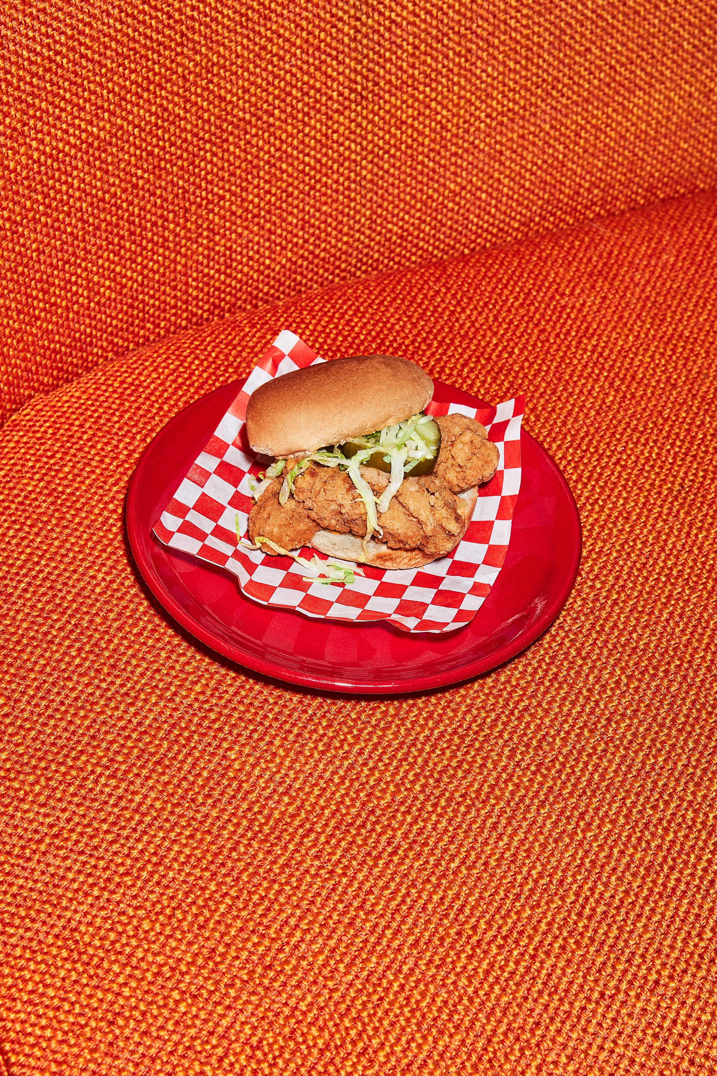 Chicken sandwich with lettuce and pickles on a red checkered paper on a red plate.