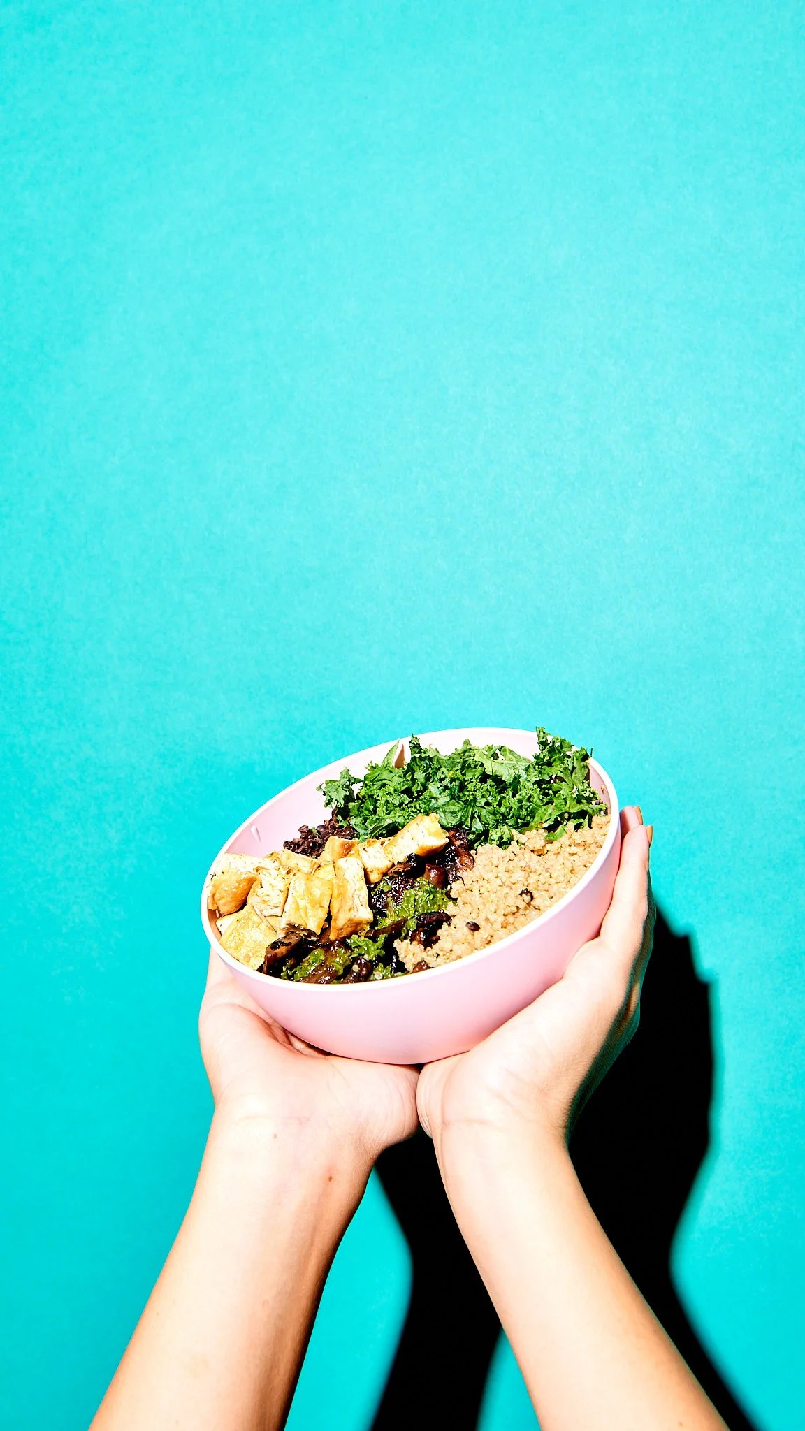 Hands holding a pink bowl filled with salad ingredients against a bright blue background.