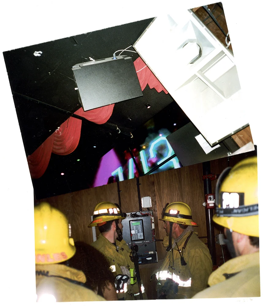 Four firefighters wearing yellow helmets and turnout gear inside a building, gathered around a control panel on the wall.