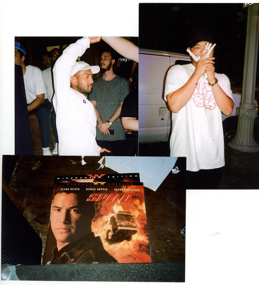 A group of people gathered indoors, including a man in a white hoodie and cap raising his hand, and another man on the right covering his mouth with his hands. On a table in the foreground, there is a movie poster for the film 'Speed' featuring a man