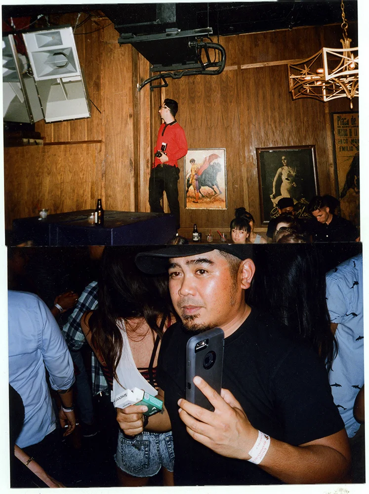 People in a crowded bar or club. One man in the foreground is taking a selfie, holding a phone and a pack of cigarettes, wearing a black shirt and a black hat. The background features wooden walls, framed artwork, and people socializing.