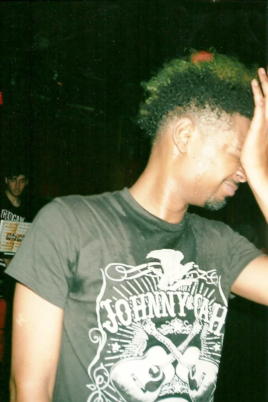 A man with green and blonde hair smiling, wearing a black T-shirt with white graphics of a guitar, eagle, and the words "Jo Hny Gah." Another person is visible in the background, looking towards the camera in a dark setting.
