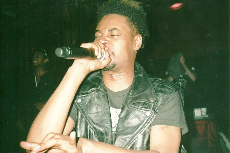 A man with curly hair and a goatee singing into a microphone, wearing a black leather vest over a black t-shirt, in a dimly lit setting.