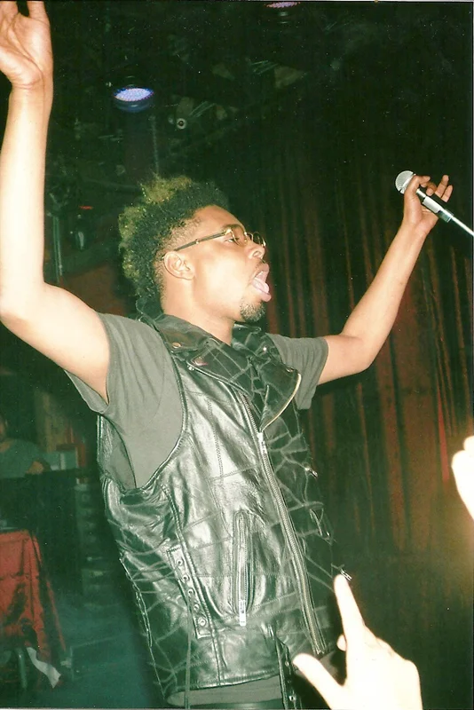 A man singing or shouting into a microphone with his arms raised, wearing glasses, a black leather vest over a gray t-shirt, in a dark indoor setting with curtains in the background.