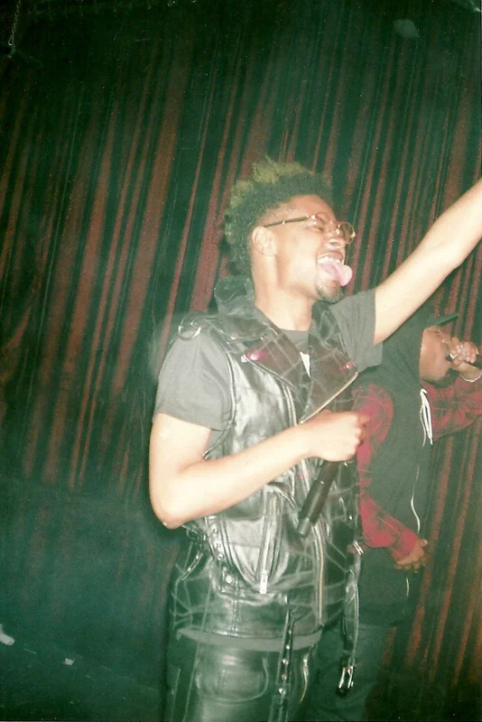 A man singing into a microphone on stage, wearing glasses, a black leather vest, and black leather pants, with a colorful curtain backdrop. music, editorial, album cover, photoshoot, conceptual, concept art