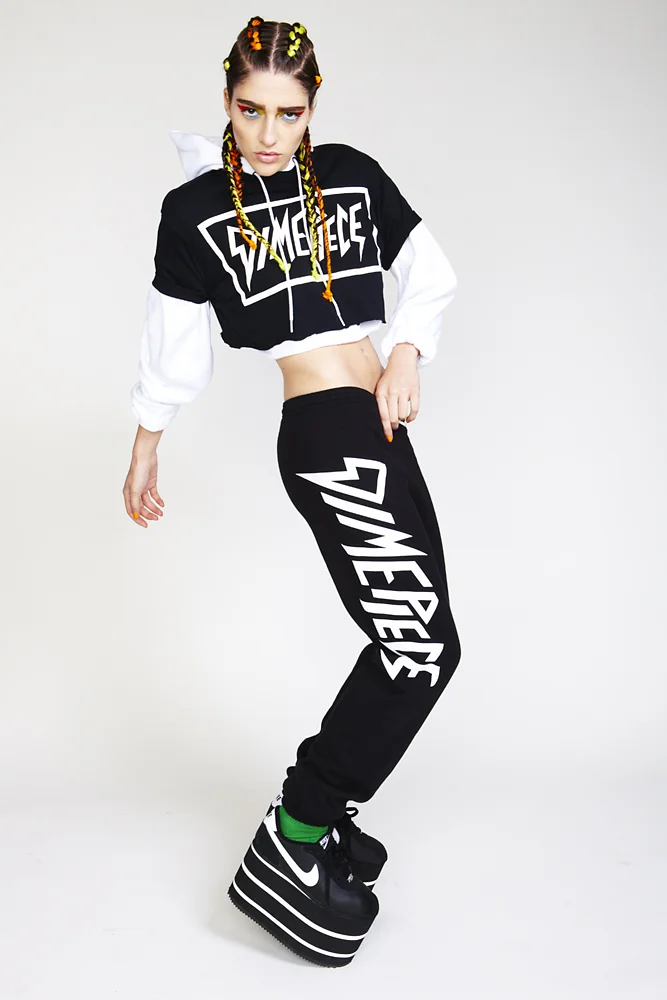 Young woman with braided hair and colorful beads, wearing a black and white crop hoodie with 'Supreme' logo, black sweatpants with 'Supreme' written vertically, and platform Nike sneakers with green socks, posing against a plain background.