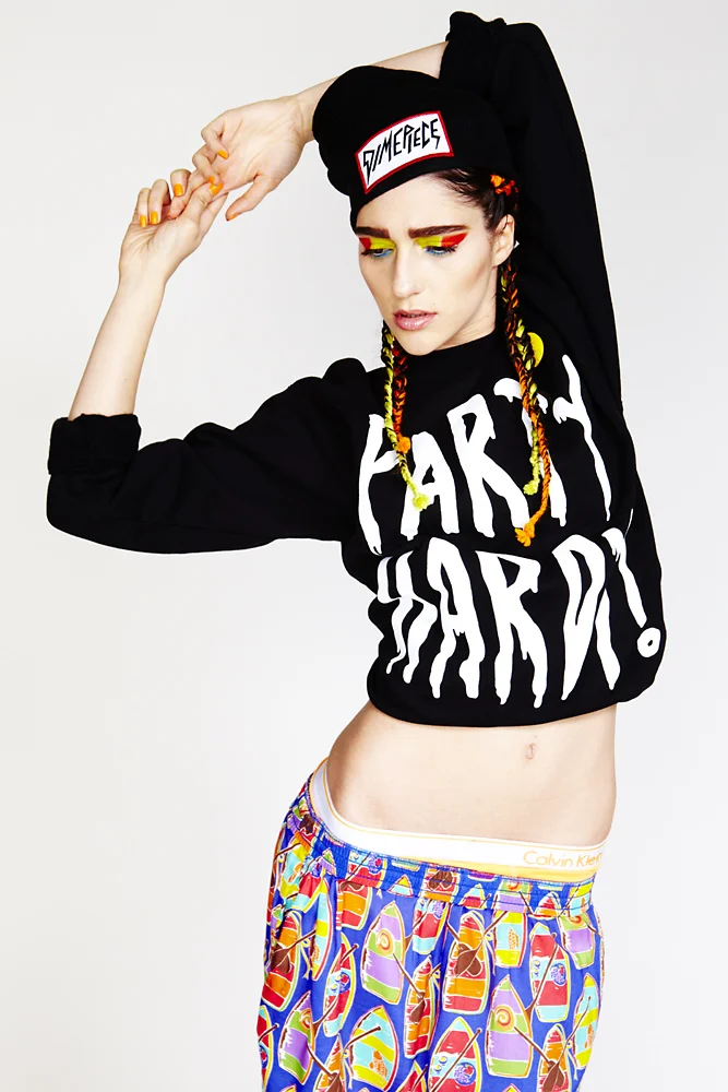 Young woman with colorful rainbow makeup, wearing a black beanie with red and white logo, from Fame Fétiche, a black oversized sweatshirt with white bold text, and patterned shorts featuring various colorful designs, posing with her right arm raised 