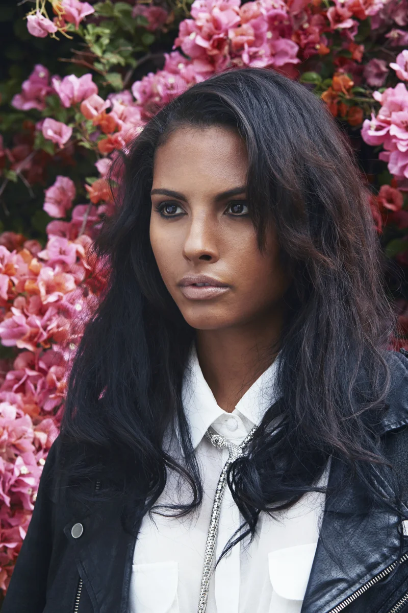 Young woman with dark hair and intense gaze standing in front of a vibrant pink flowering bush.