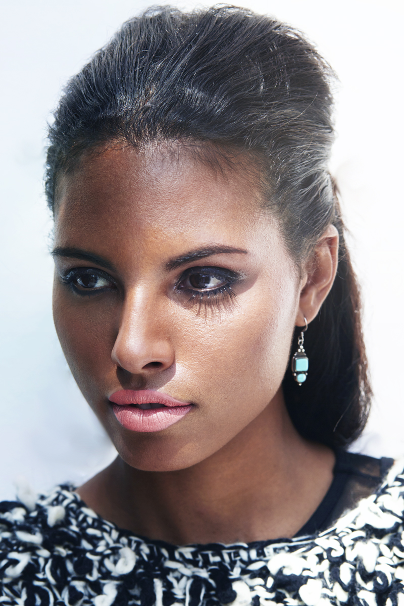 Close-up portrait of a woman with dark hair styled back, wearing makeup with dark eye makeup and pink lipstick, and a black and white patterned top with turquoise earrings.