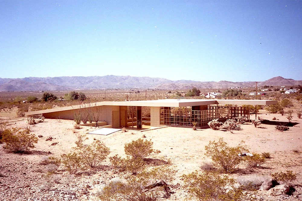A modern, flat-roofed house in a desert landscape with mountain view in the background, surrounded by shrubs and sparse desert vegetation.