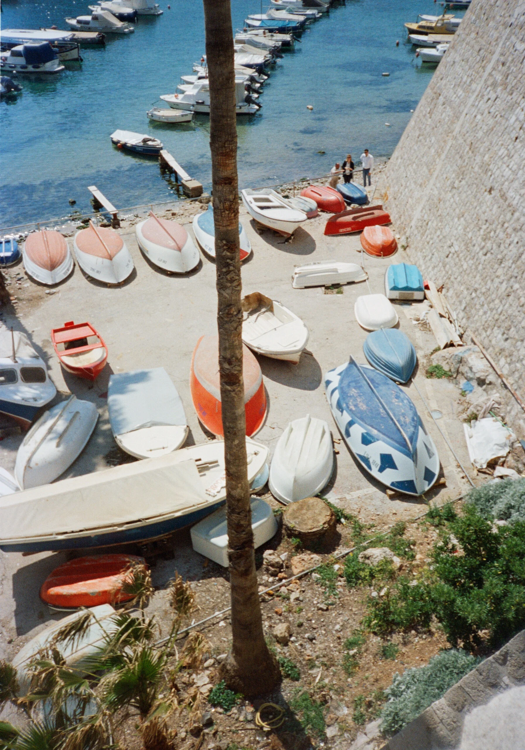 A small boat yard with various boats on land, some covered and some uncovered, next to a stone wall and a waterfront. A couple of people are walking near the water.