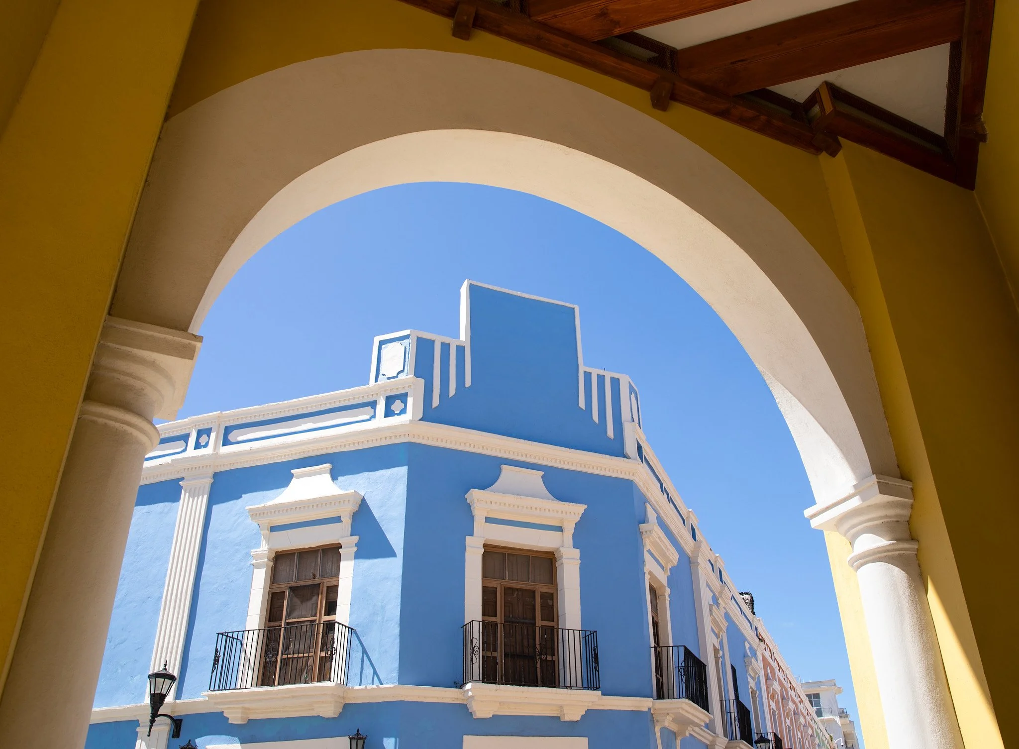 View of a blue building with white architectural details, framed by a yellow arch and wooden roof overhang, under a clear blue sky.