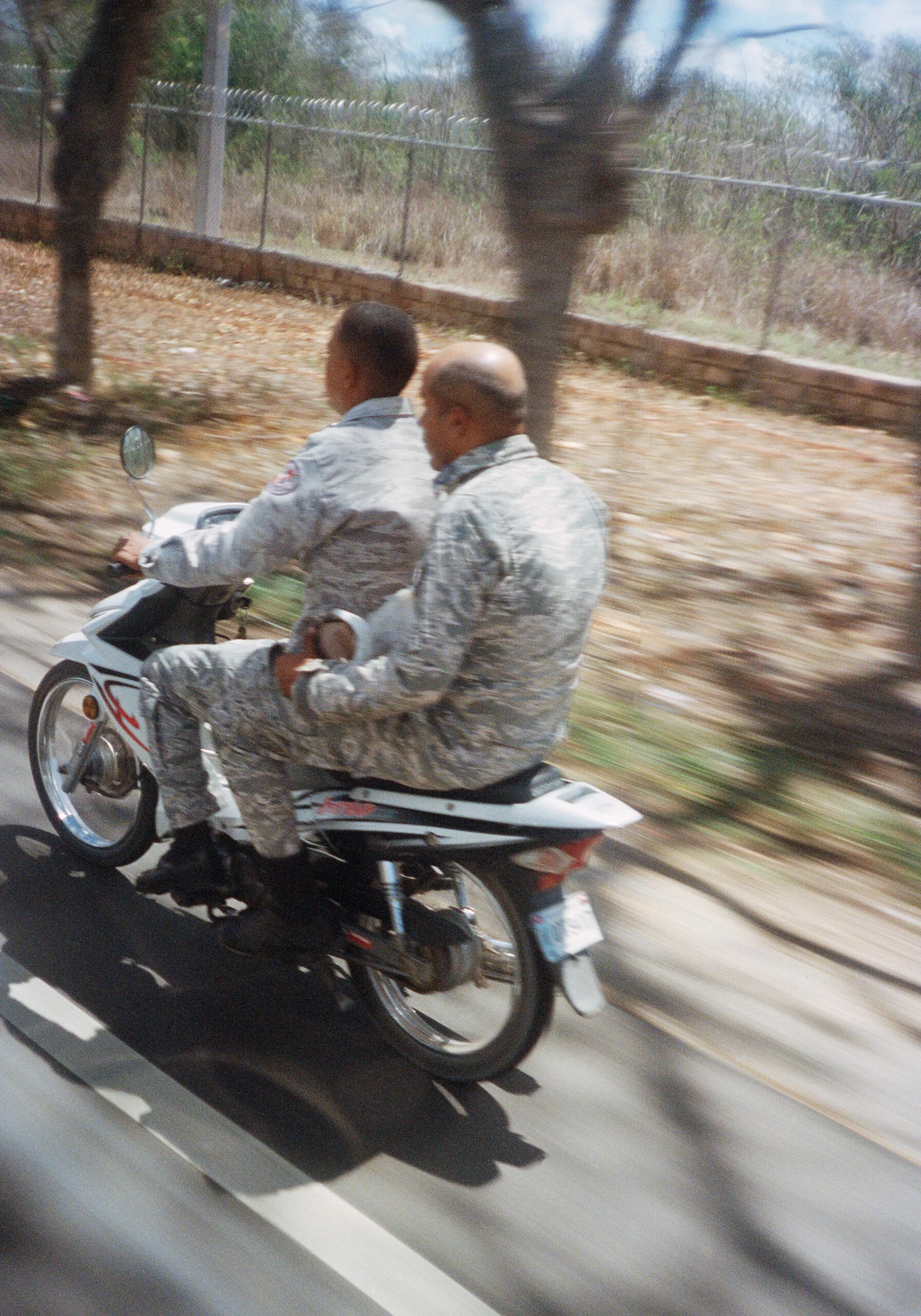 Two men in military uniforms riding a motorcycle on a road with a barbed wire fence and trees in the background.