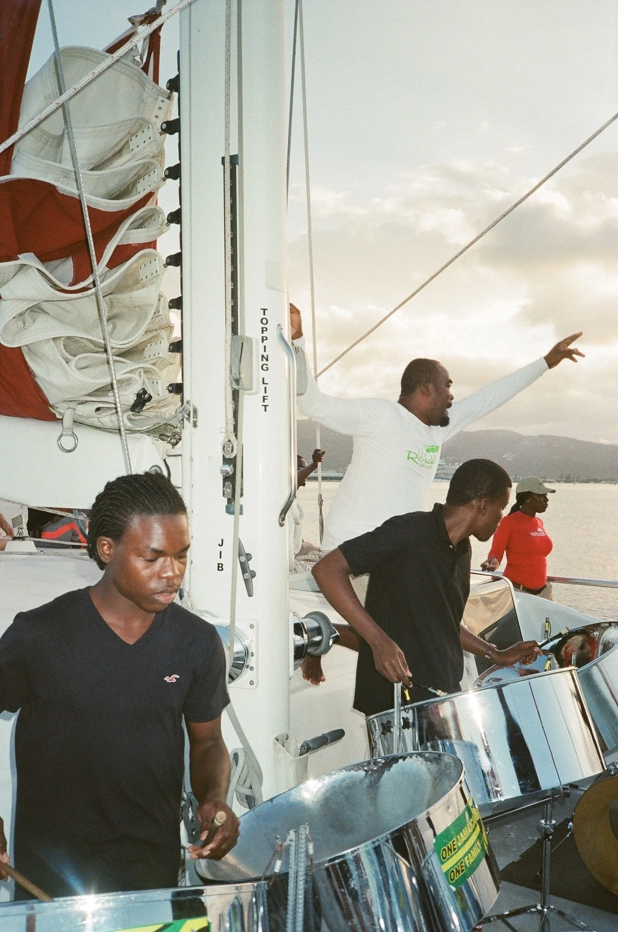 Musicians playing steel drums on a boat during sunset, with mountains and water in the background.