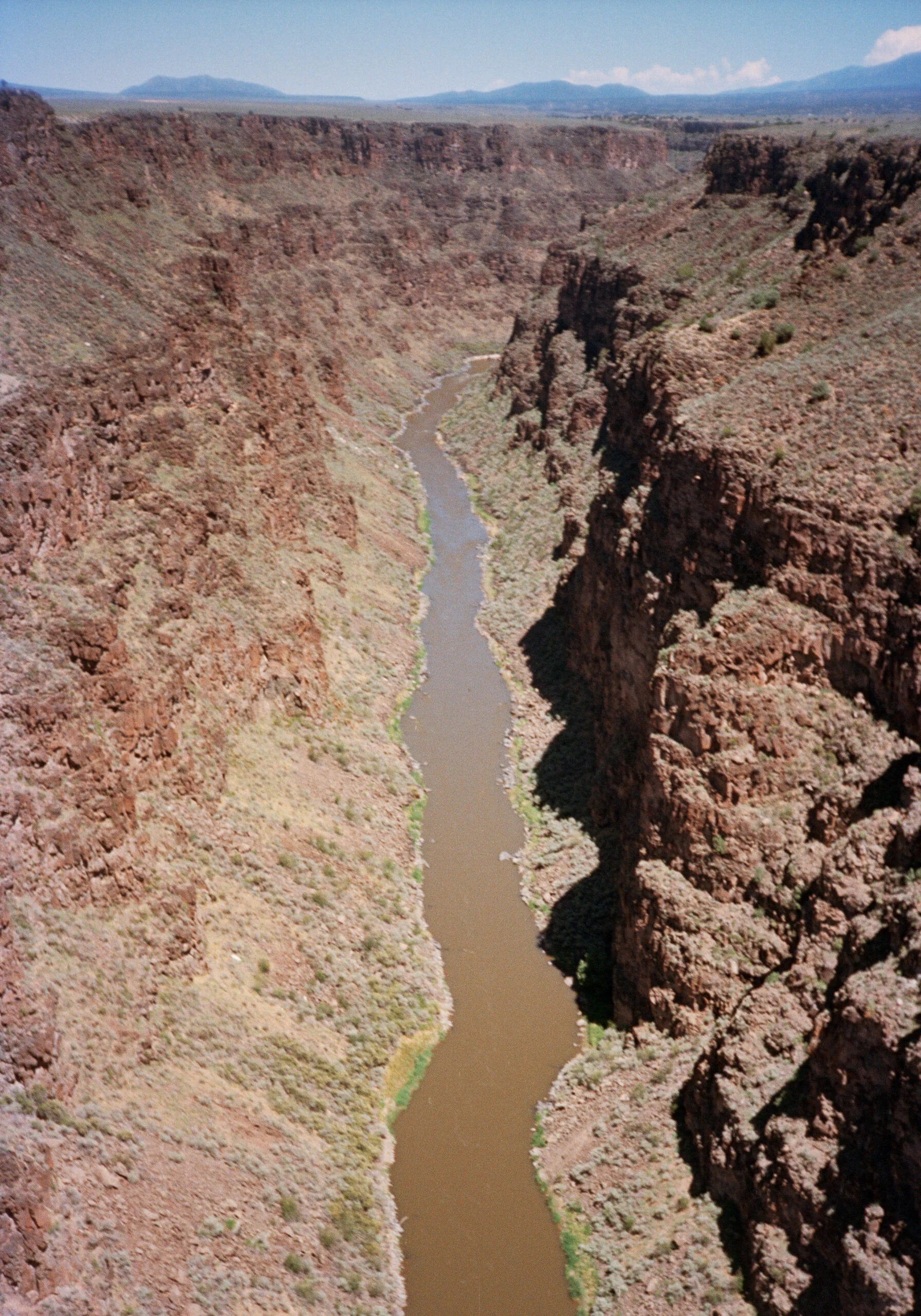 A river flows through a deep, narrow canyon with steep, rugged red rock walls on both sides, under a partly cloudy sky.