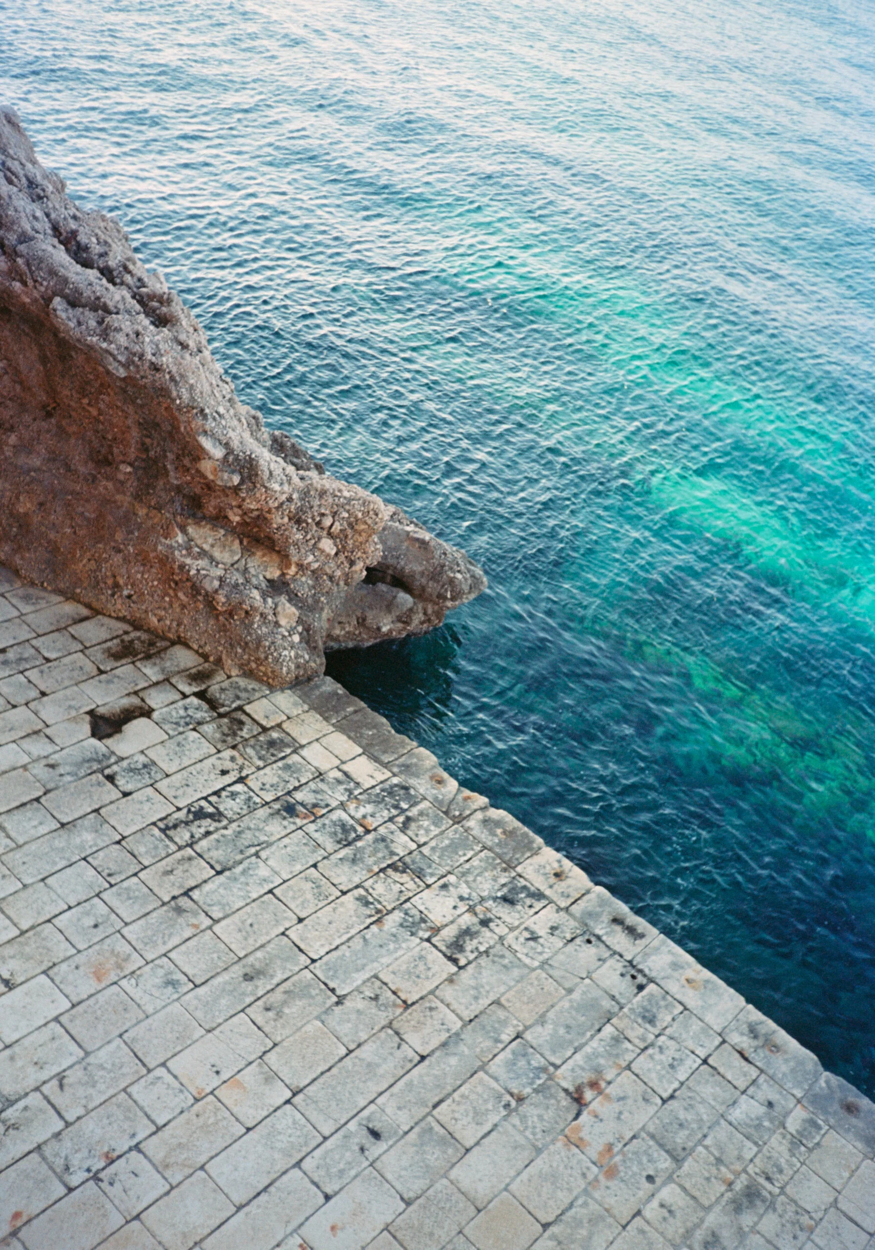 View of a rocky cliffside meeting the ocean with blue and green water, next to a paved stone walkway.