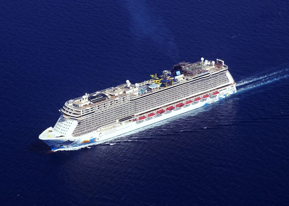 A large cruise ship sailing on the open ocean, with multiple decks and red lifeboats visible along the side.