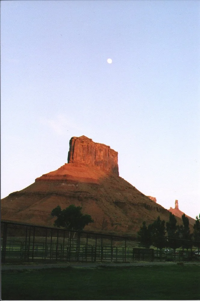 A large rocky formation or mesa with a flat top, illuminated by sunlight, under a clear sky with visible moon.