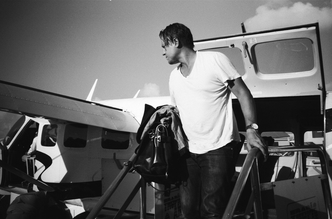 Man walking up the stairs to an airplane on a tarmac, holding a bag and wearing casual clothing in black and white.