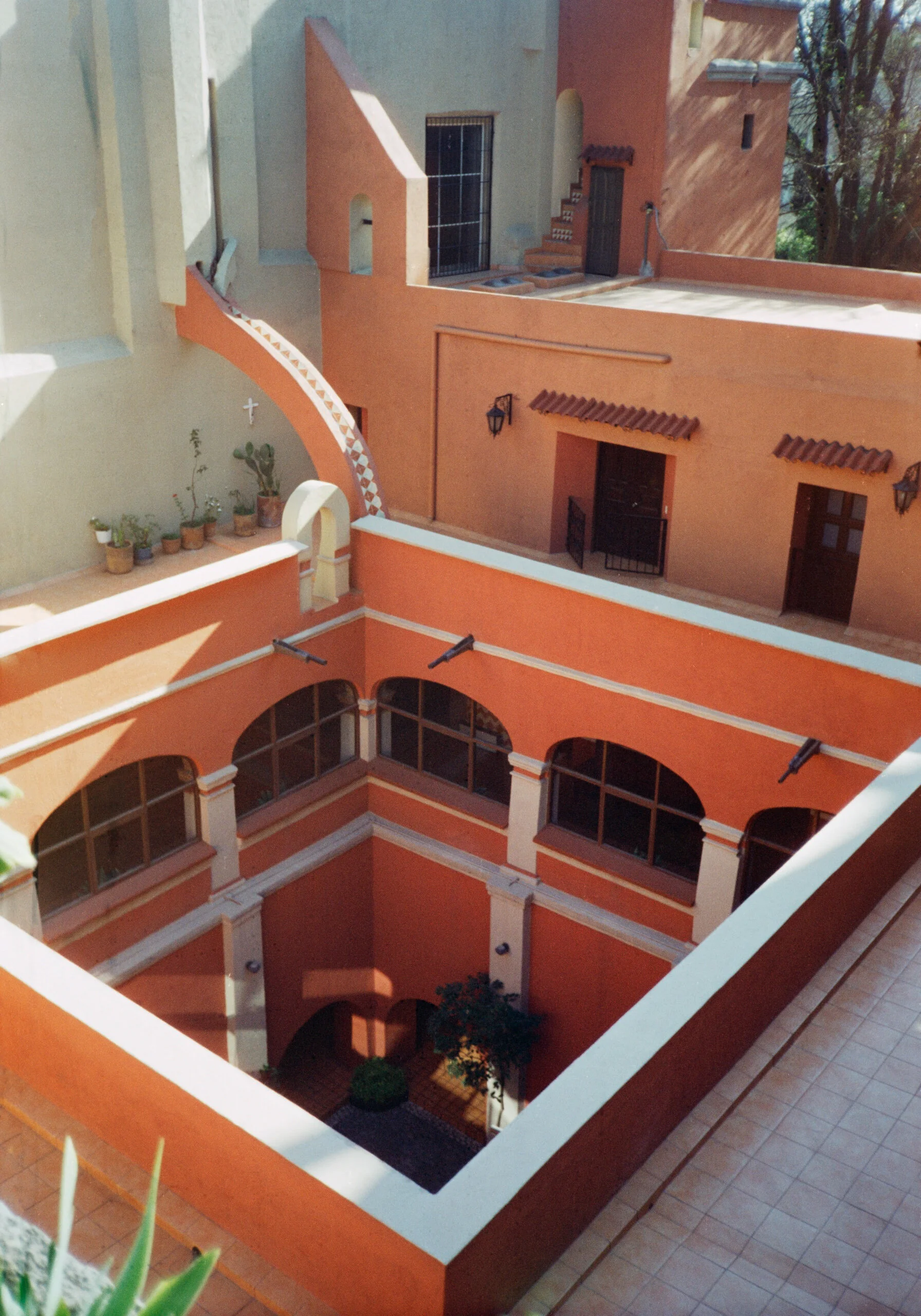 Photo of a multi-story courtyard with orange and beige walls, arched windows, potted plants, small trees, and outdoor lighting fixtures.