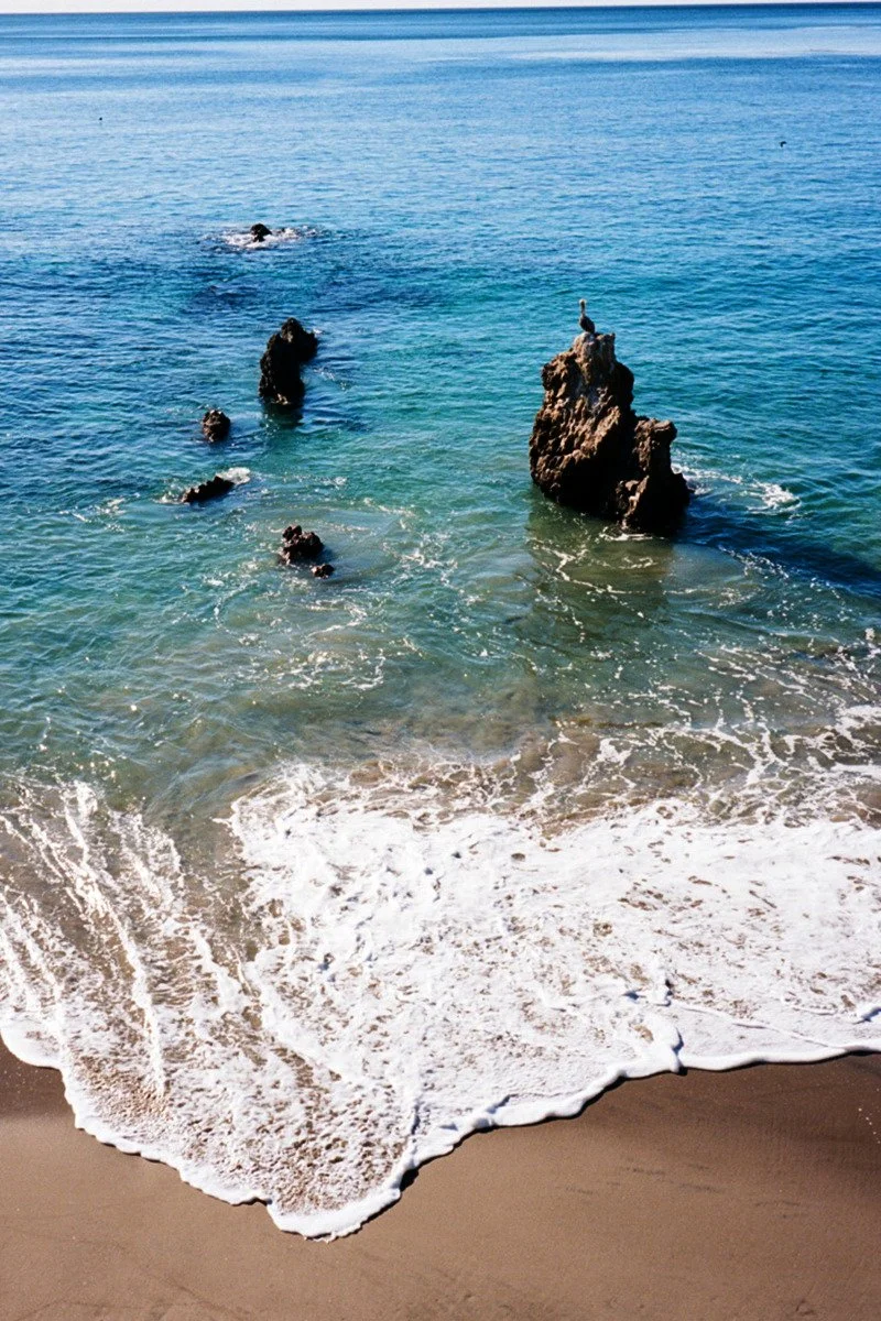 Sea rocks with a bird perched on top, waves crashing on the sandy beach, and the ocean stretching to the horizon under a clear sky.