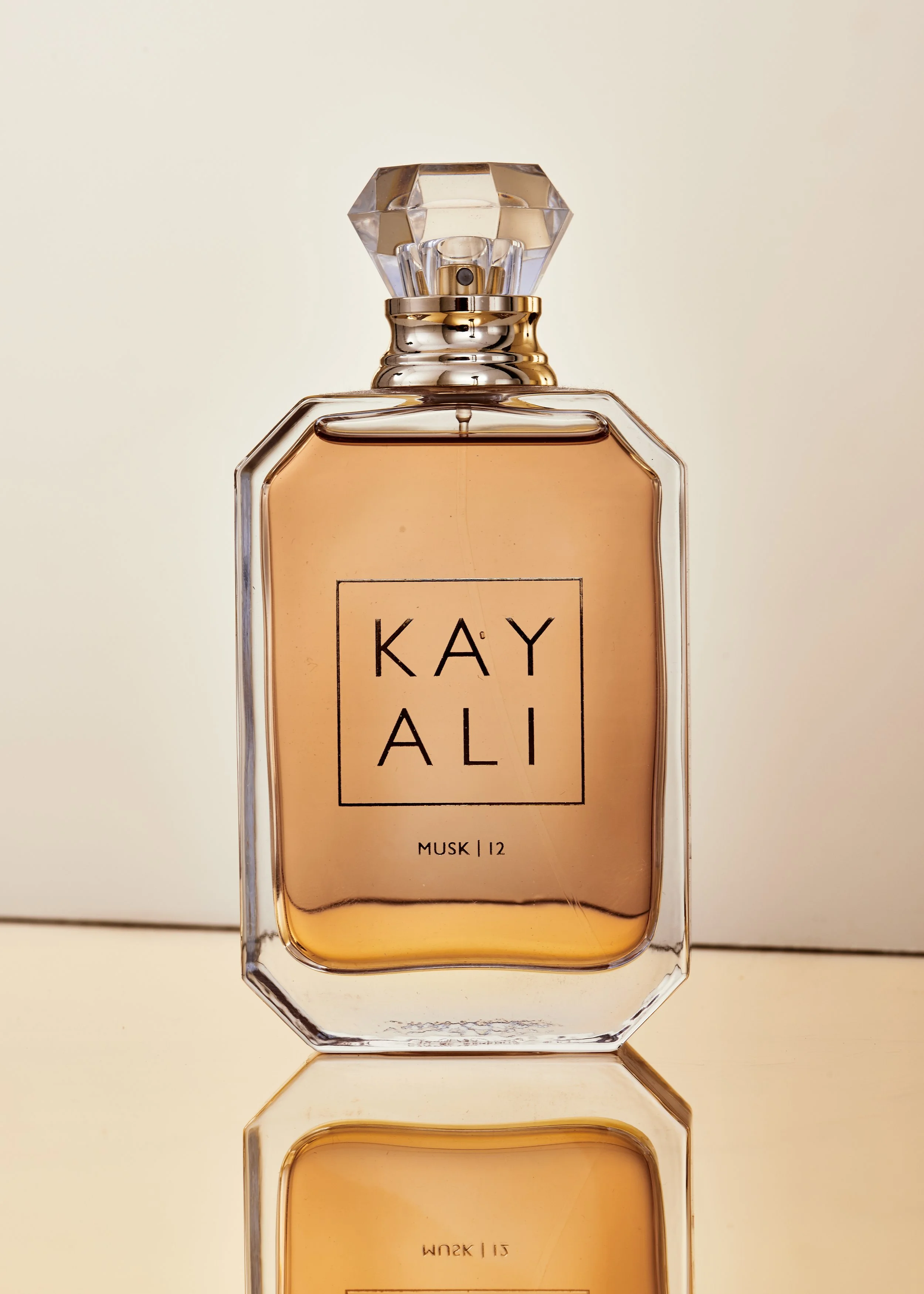 A glass perfume bottle with a rectangular shape, labeled Kayali Musk 12, filled with a peach-colored fragrance, reflecting on a surface.