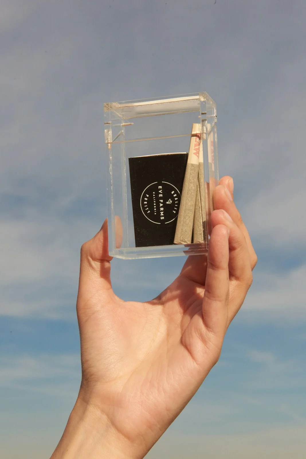 A clear acrylic box being held up against a blue sky with some clouds. Inside the box, there is a black card with white text that reads 'EYE FARMS' and some small rolled-up paper or cardboard items.