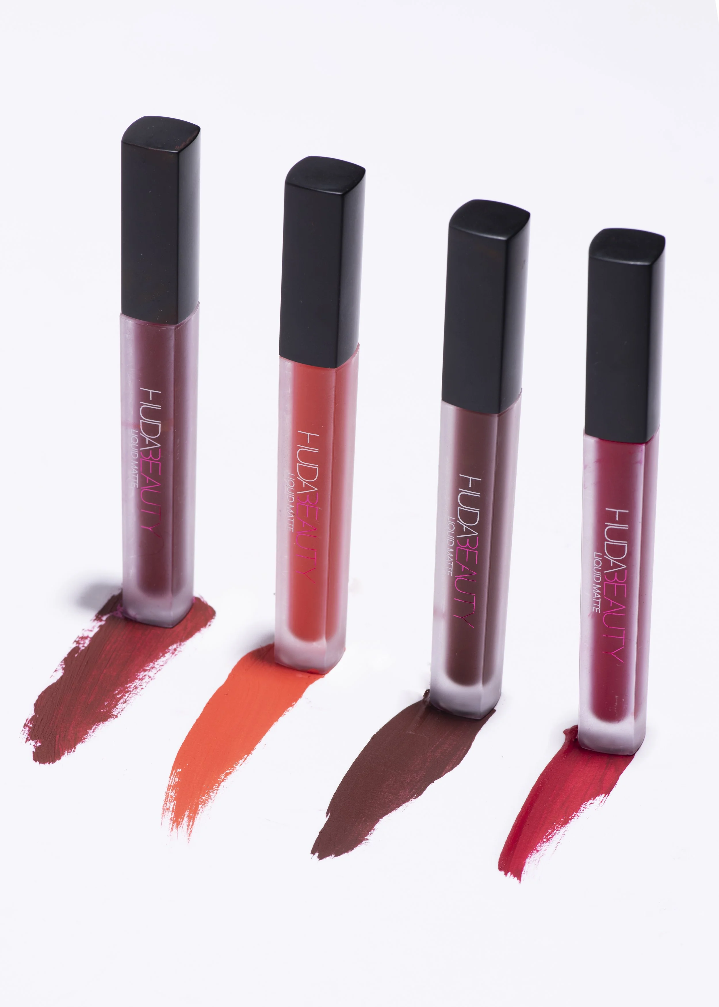 Four lip gloss tubes from Huda Beauty standing upright with colored swatches of their shades beneath each, on a white background.