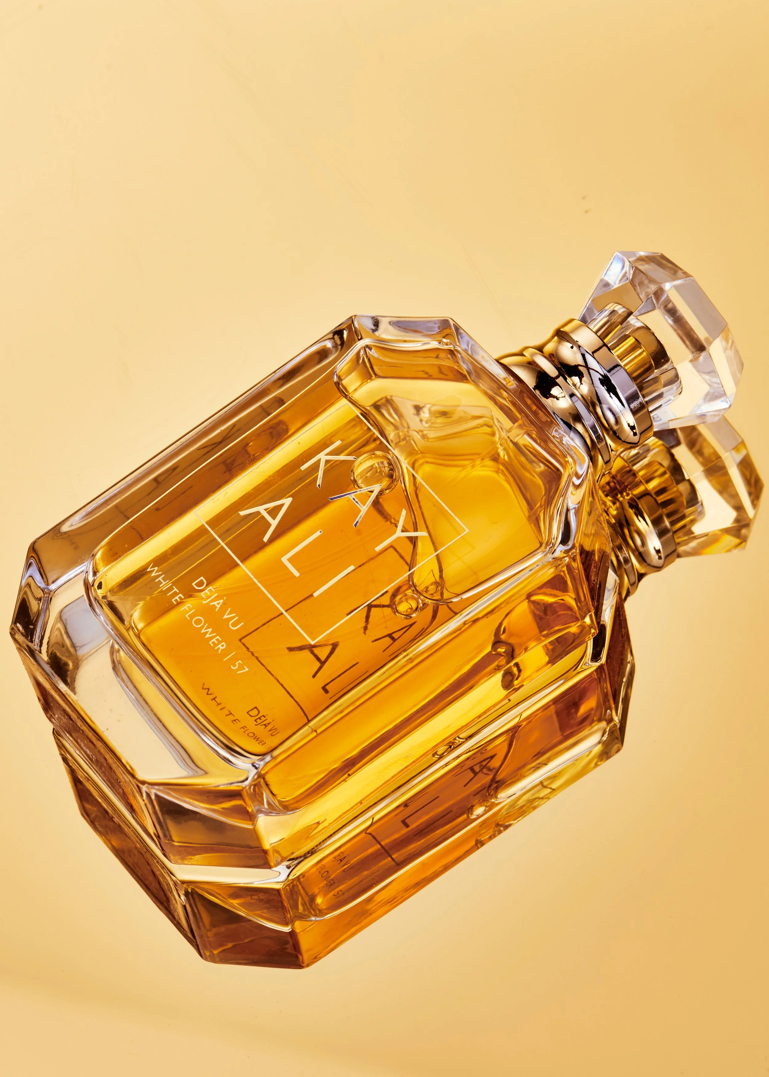 A close-up of a designer perfume bottle labeled 'KAYALI' with the scent 'Deja Vu White Flower 57,' resting on a reflective yellow surface.