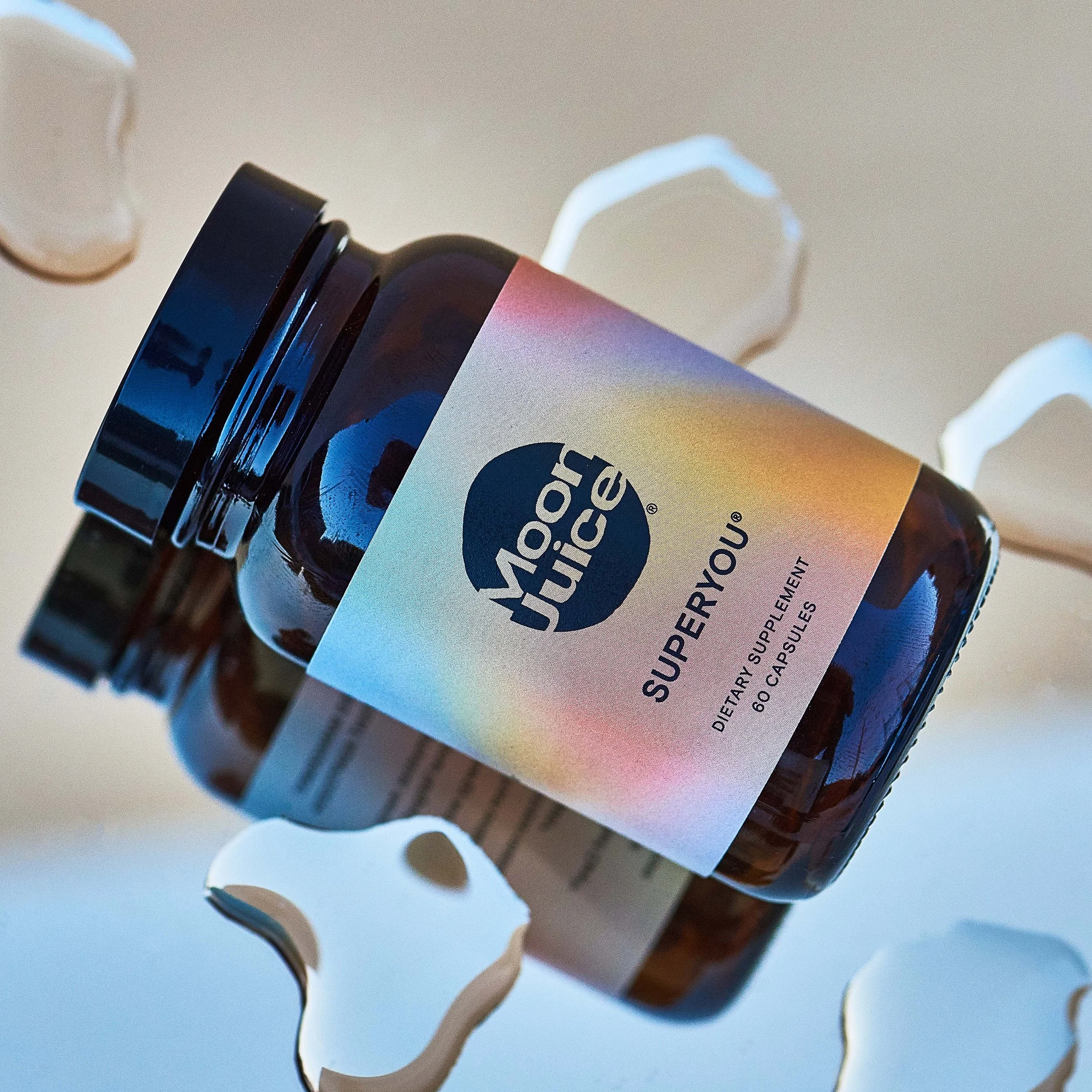 A brown bottle of Nourish Your Superfood dietary supplement with a pastel rainbow label, surrounded by curly paper shreds on a reflective surface.
