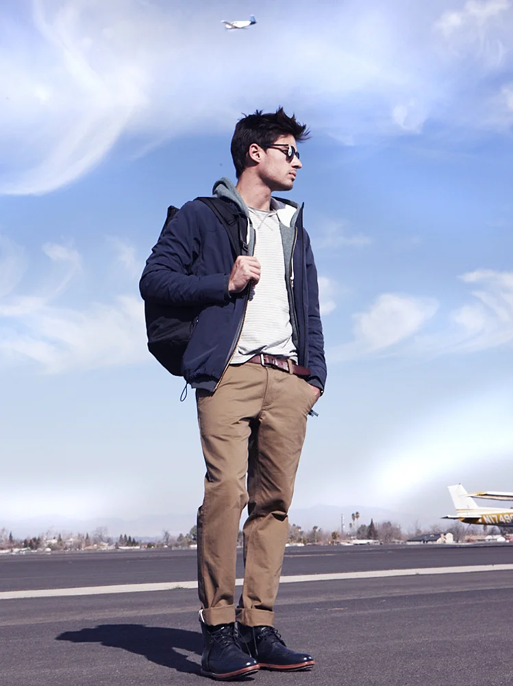 A man standing on an airport runway with a backpack, wearing sunglasses, a navy jacket, gray hoodie, striped shirt, khaki pants, and black boots. An airplane is flying overhead against a blue sky with some clouds.