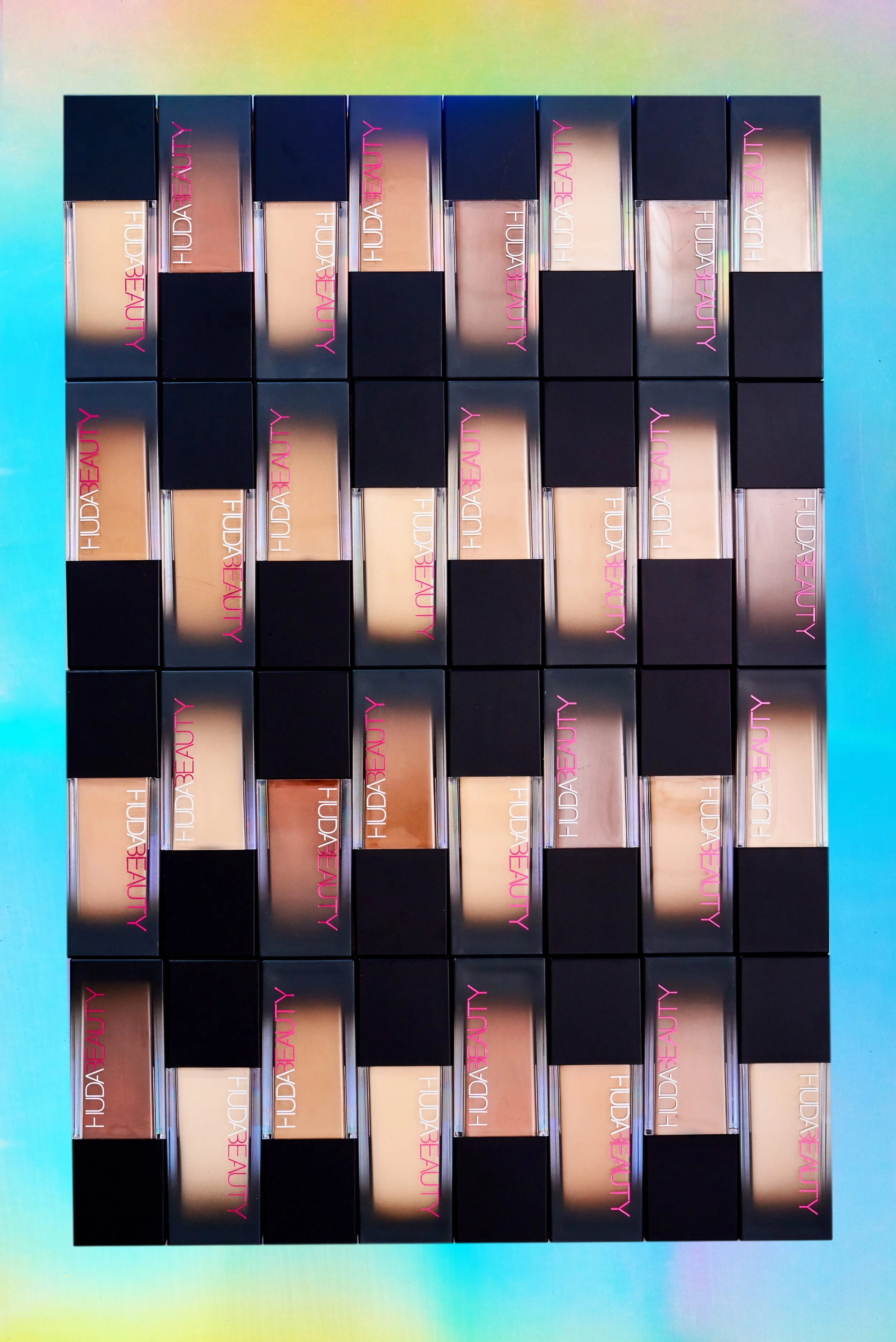 Multiple Huda Beauty makeup concealer boxes arranged in a grid on an iridescent surface.