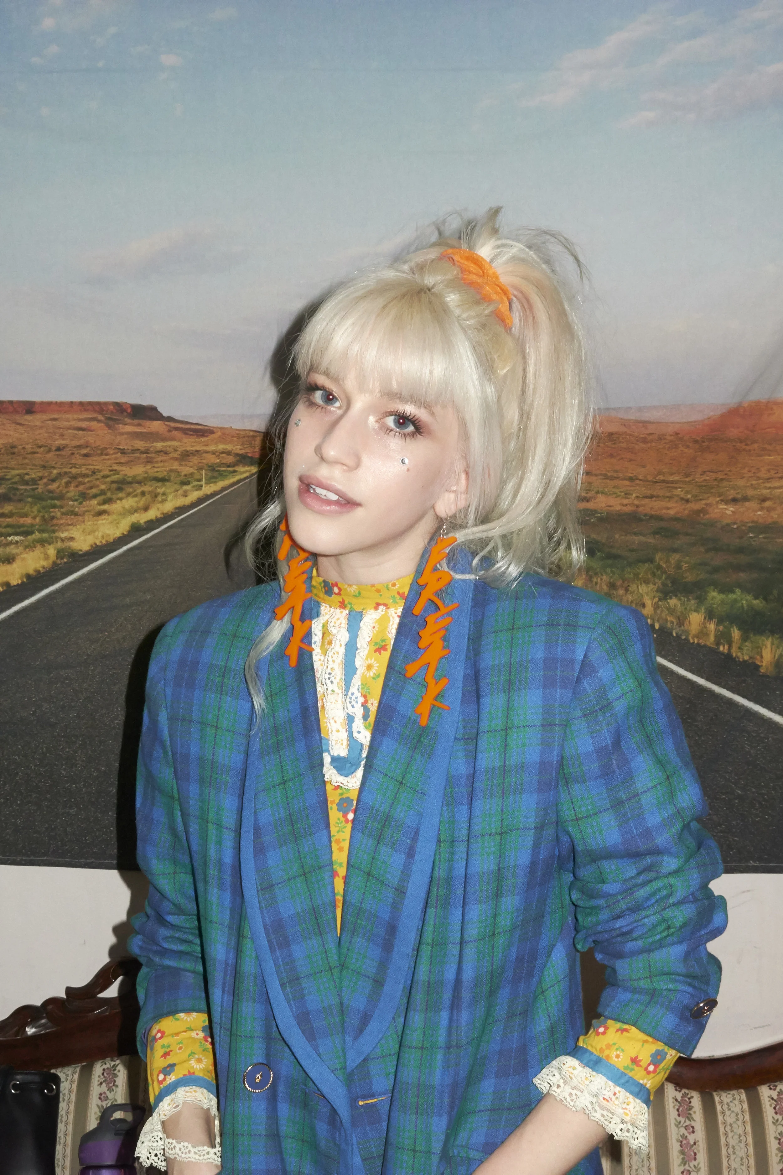 A woman with platinum blonde hair styled in a high ponytail with an orange hair tie, wearing a blue plaid blazer, yellow floral shirt, and orange earrings that spell 'FREAK'. She has diamond face gems near her eyes and is standing in front of a backd