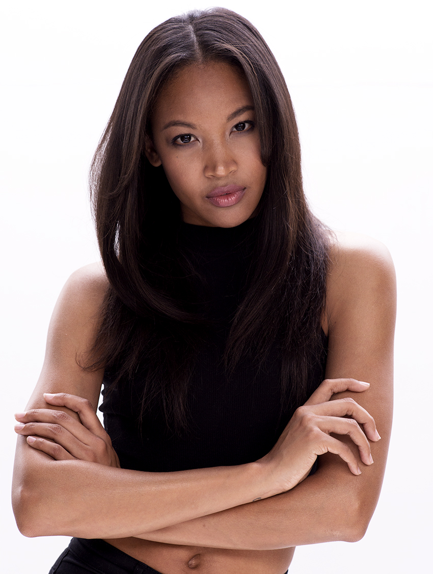 Portrait of a young woman with long dark hair, wearing a black sleeveless top, crossing her arms, looking directly at the camera against a plain white background.